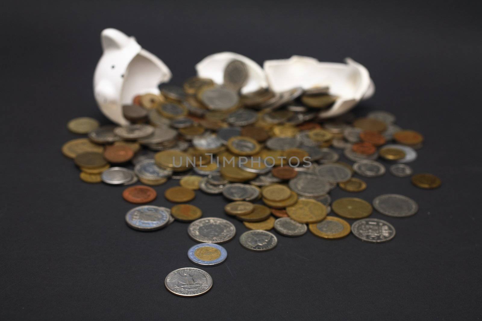 A broken piggy bank isolated on a dark background with loads of coins from around the world.