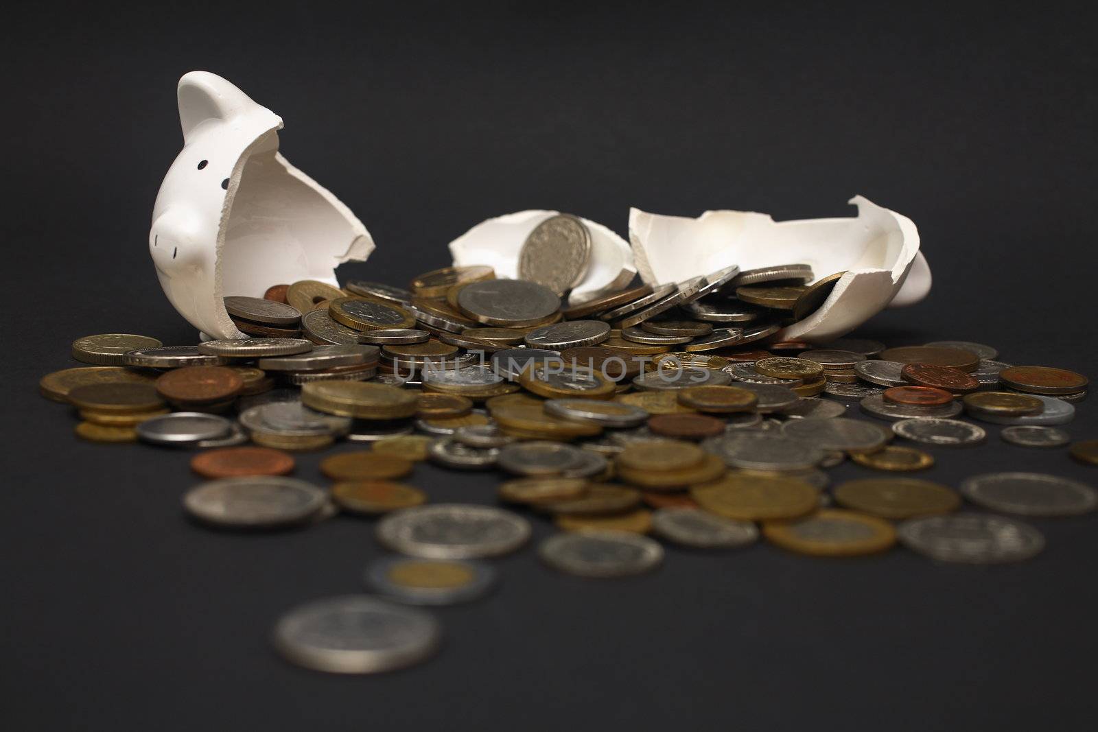 A broken piggy bank isolated on a dark background with loads of coins from around the world.