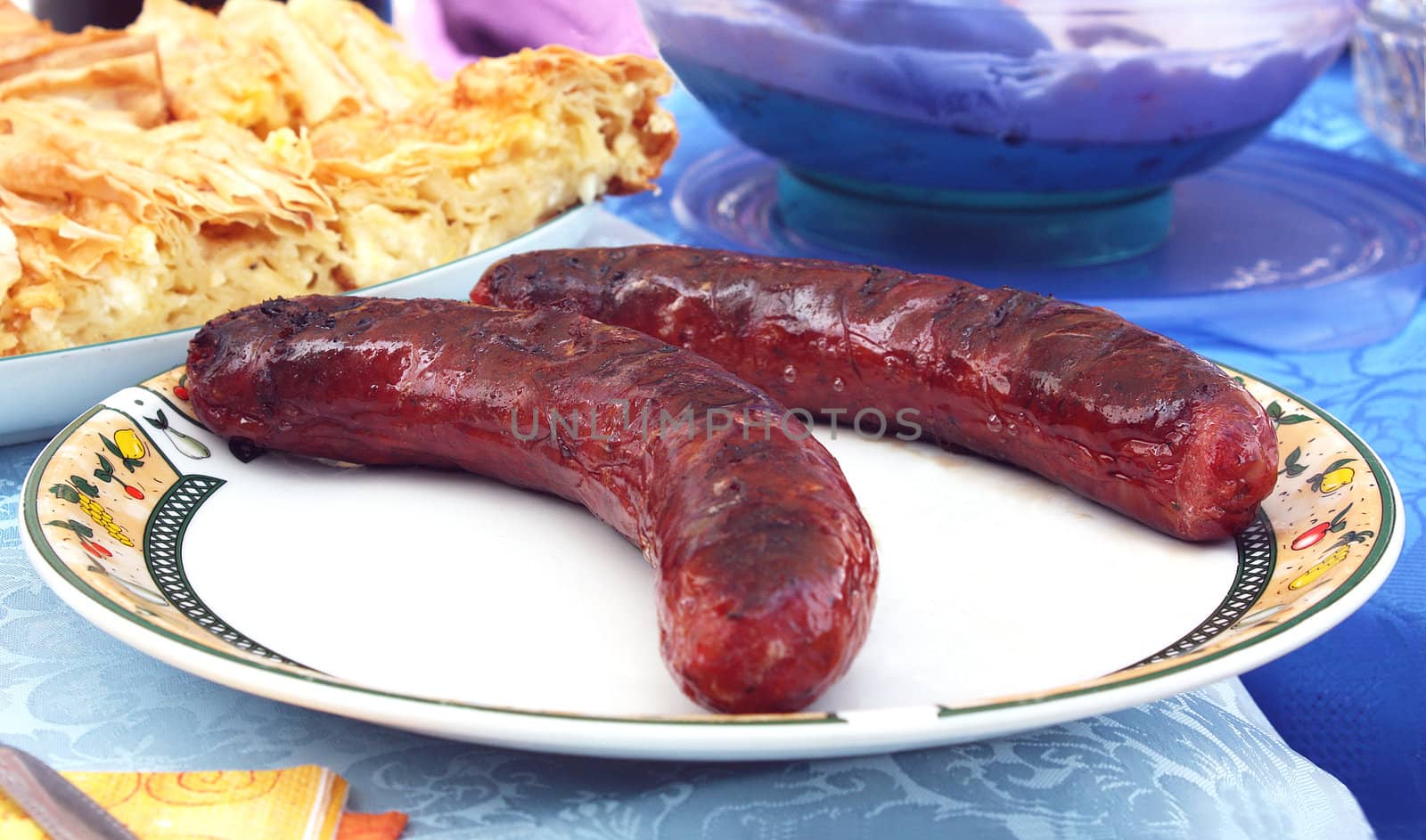 Close up of two Greek sausages on a plate. In the background there are some tiropita pieces (Greek cheese pastry).