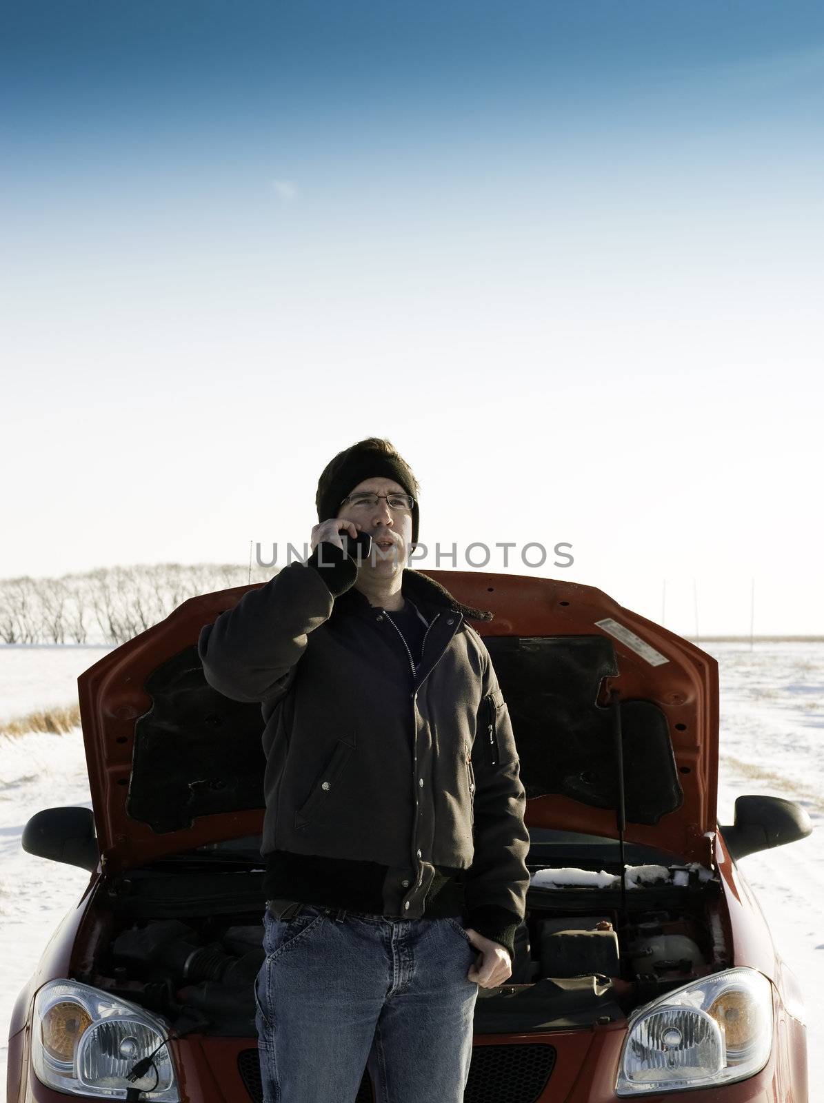A young man is calling for help on his cell phone, while standing in front of a red car with it's hood up.  Copyspace is available on the top half of the image.