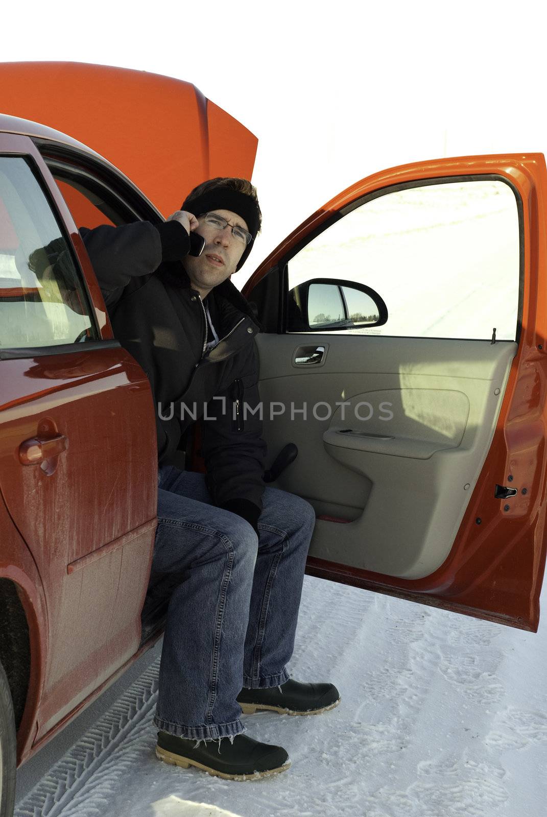 A young man using his cell phone to call for help while waiting in his car with the hood up.