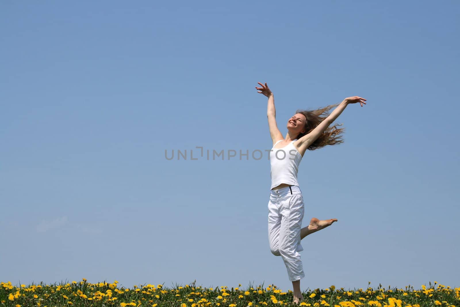 Smiling girl in summer white clothes dancing in a flowering dandelion field.