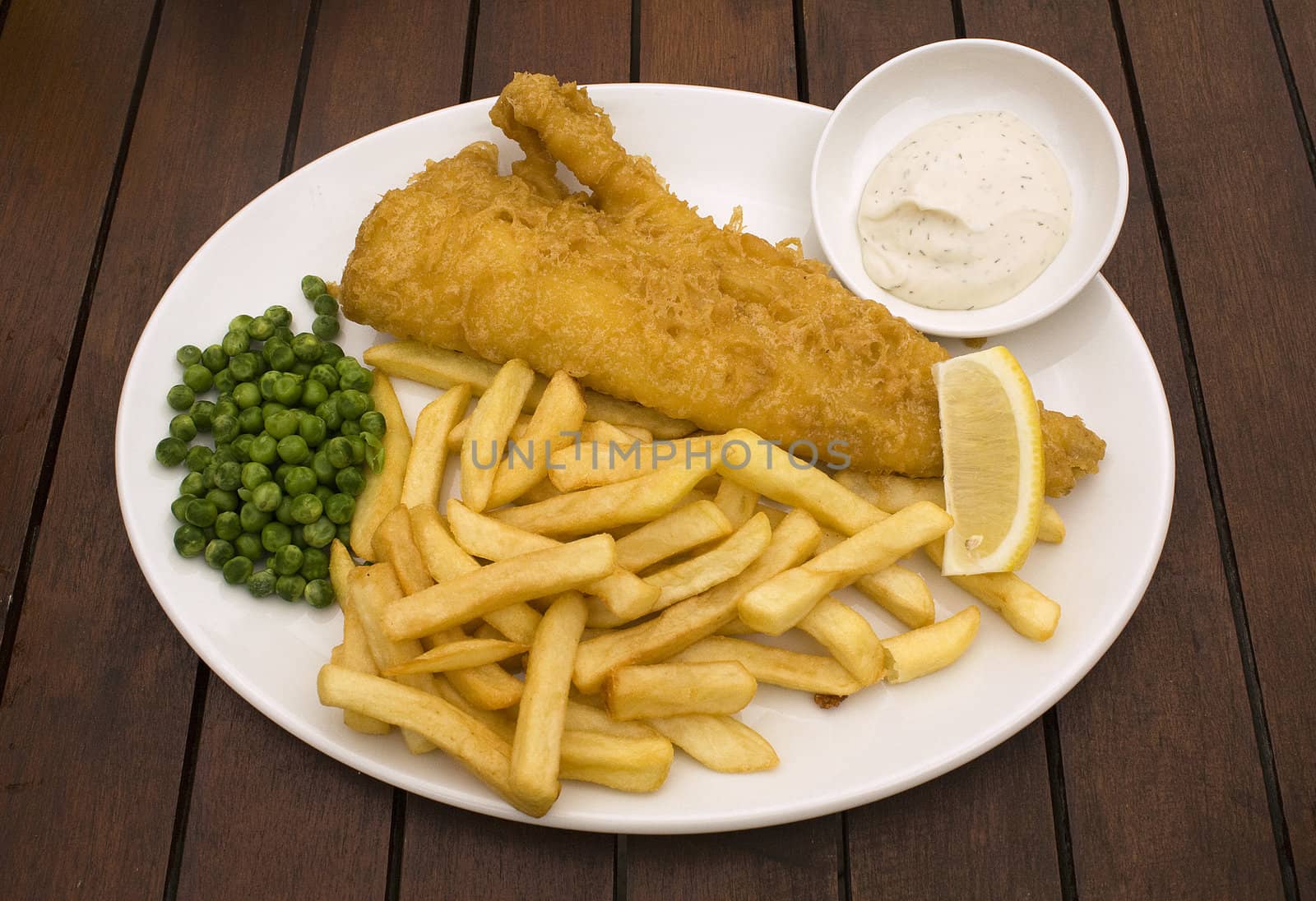 Fish and chips served with peas and tartar sauce on white plate on wooden pub table.