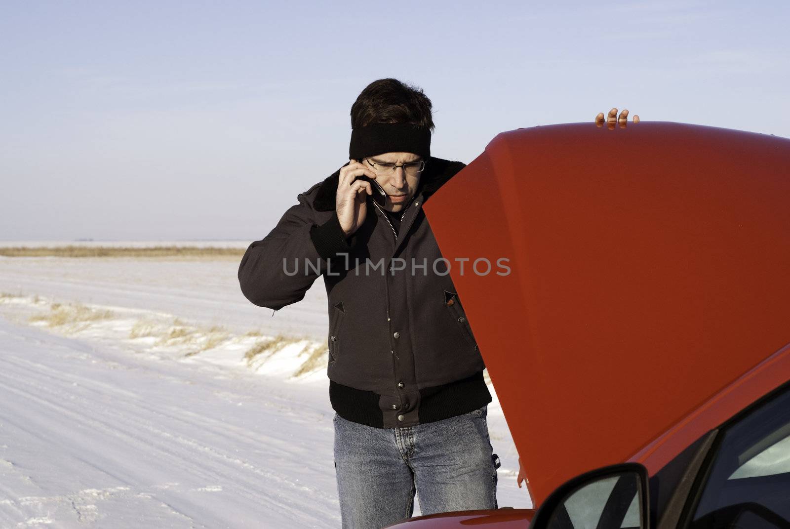 A young man is checking under the car hood while talking on a cell phone.
