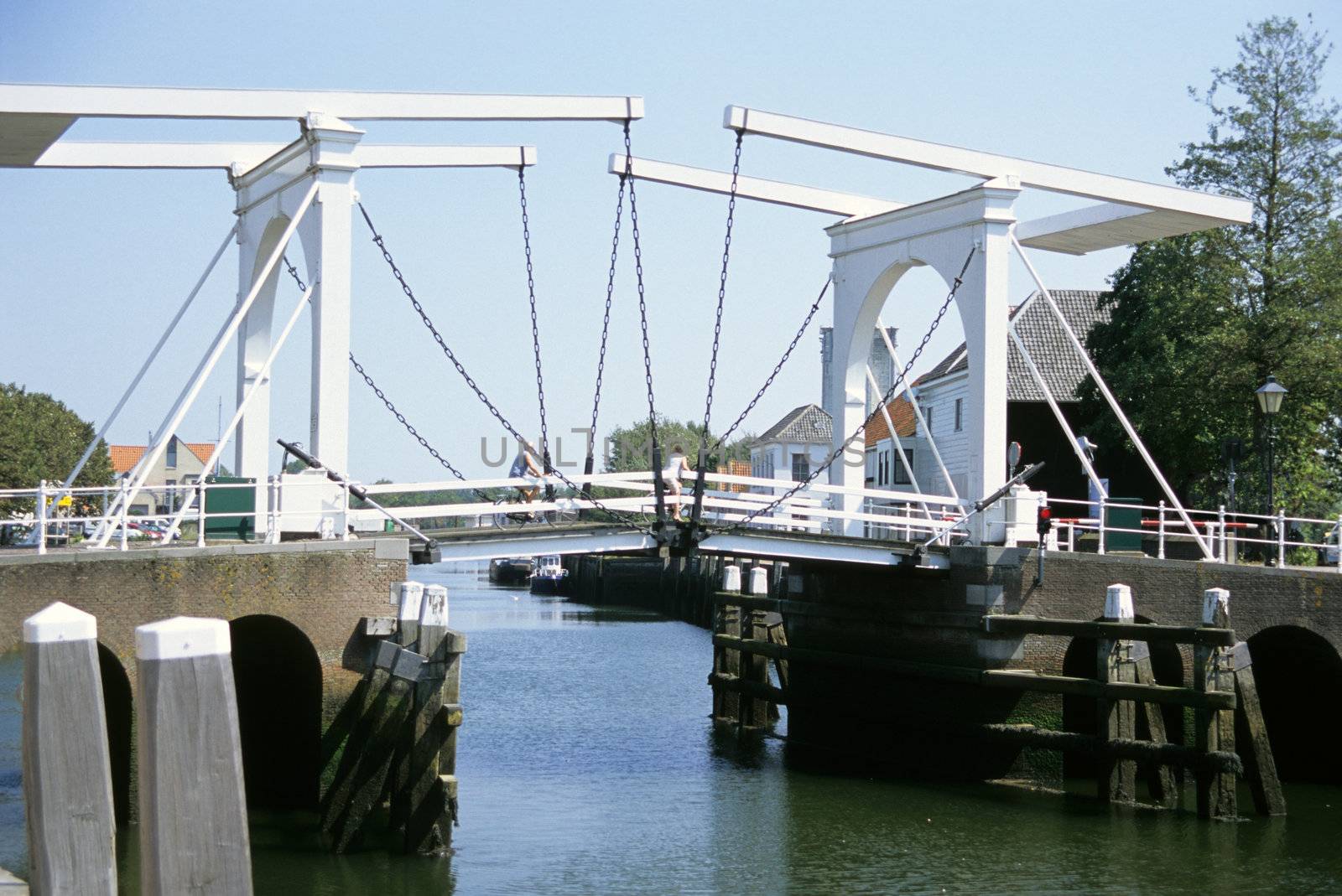 Two people ride bicycles over a traditional wooden drawbridge that spans a canal in the Netherlands.