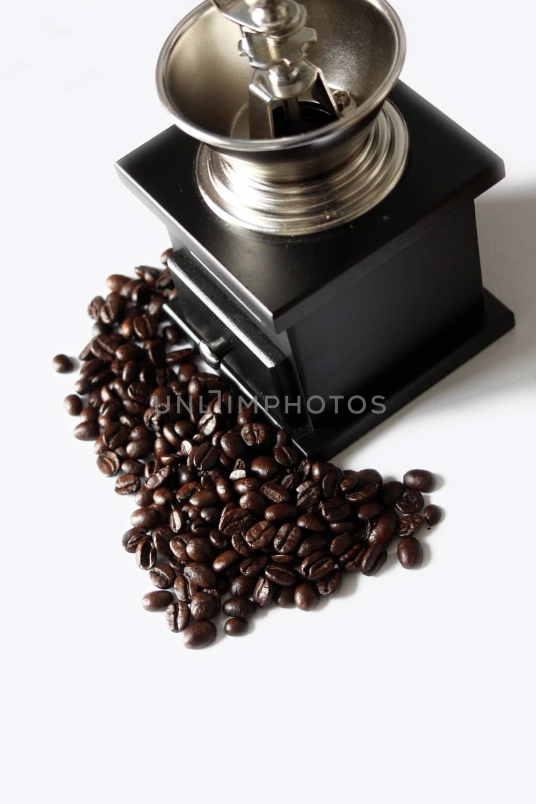A portrait format image of a black wooden hand coffee grinder set amongst a pile of coffee beans, set on a white isolated background.