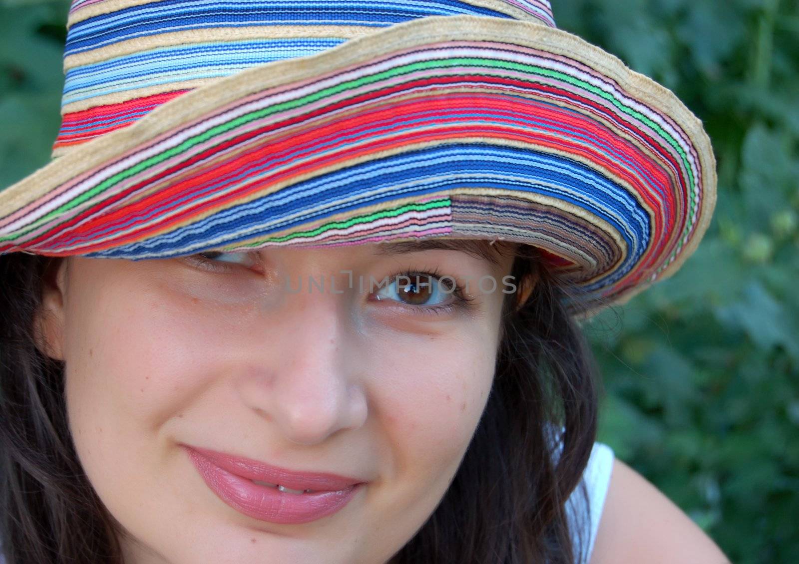 close-up of attractive young brunette in colorful hat against green leaves in the garden