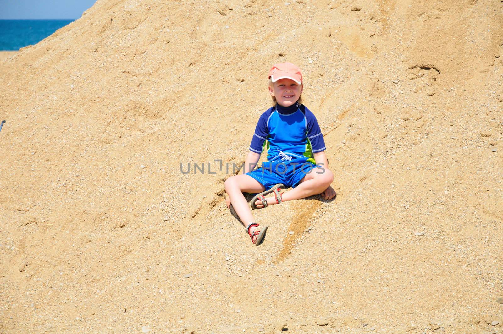 happy little blond boy wearing surfing suit is sitting in the huge pile of sand on the beach, good weather, summer is here! 