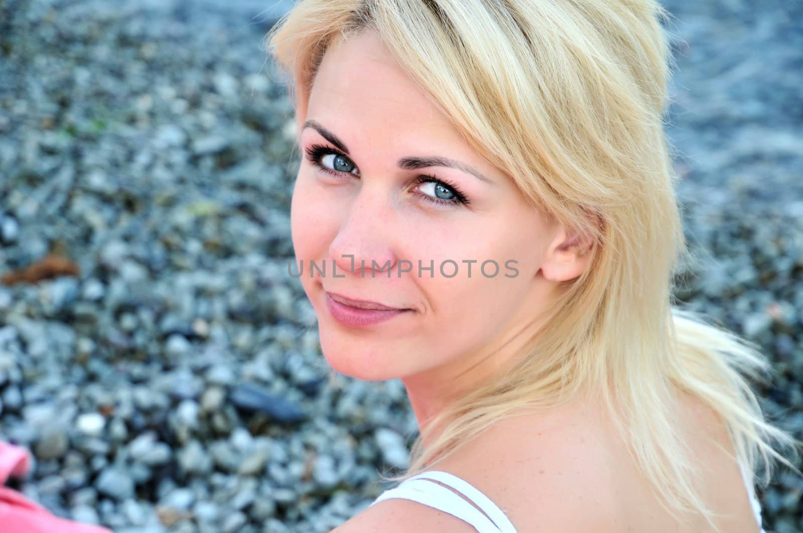 portrait of  beautiful young blond woman with stunning blue eyes, she is sitting on the 	 pebble near the sea.