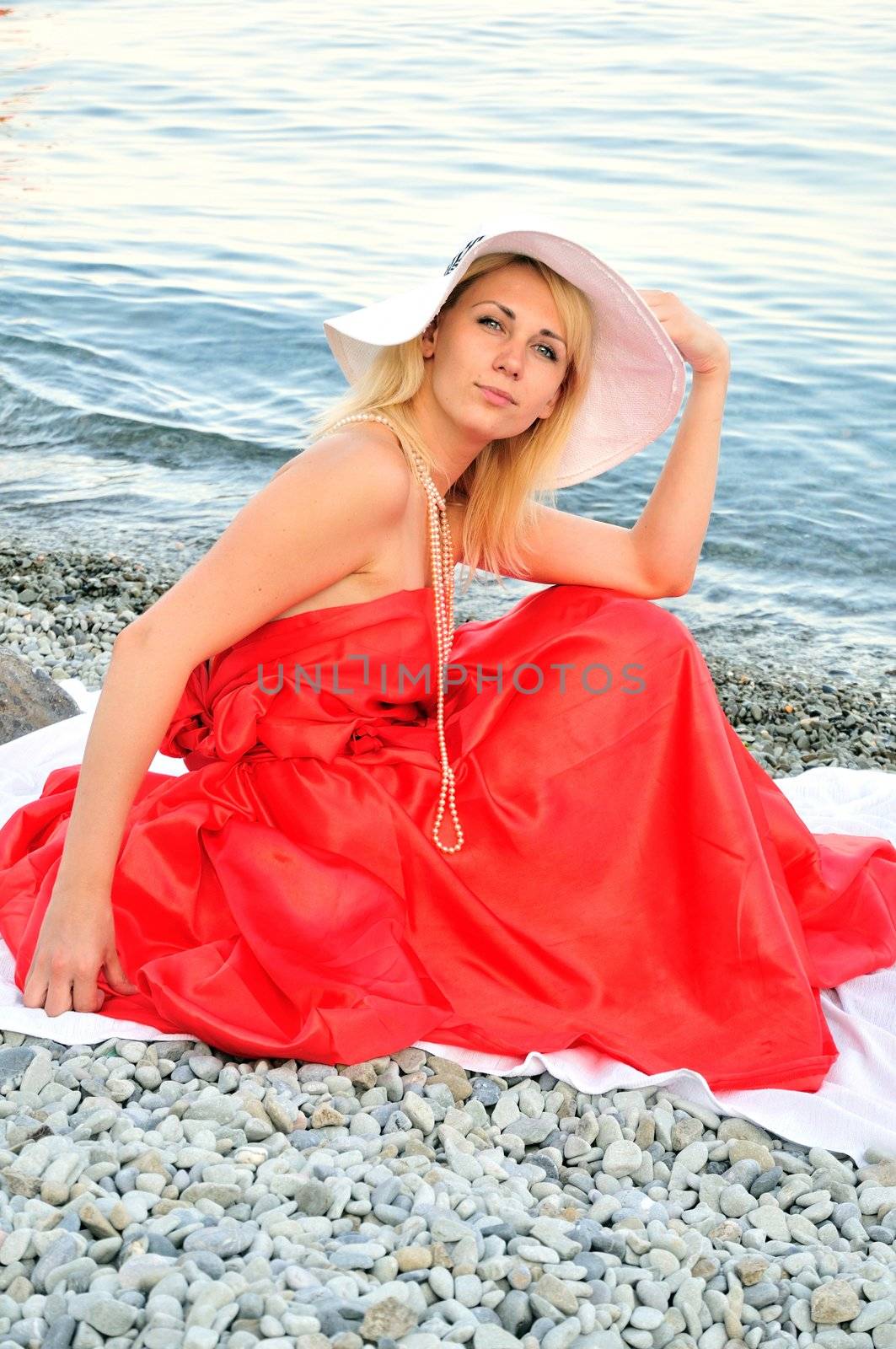 beautiful young woman wearing white hat, strand of pearls and red dress is sitting on the beach near the sea