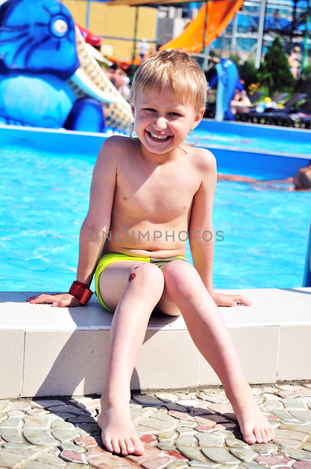 boy wearing swimming trunks is sitting close to swimming pool. summer. sunny day, boy is happy, but his kneel is already  wounded, but he is boy, so it is not unusual