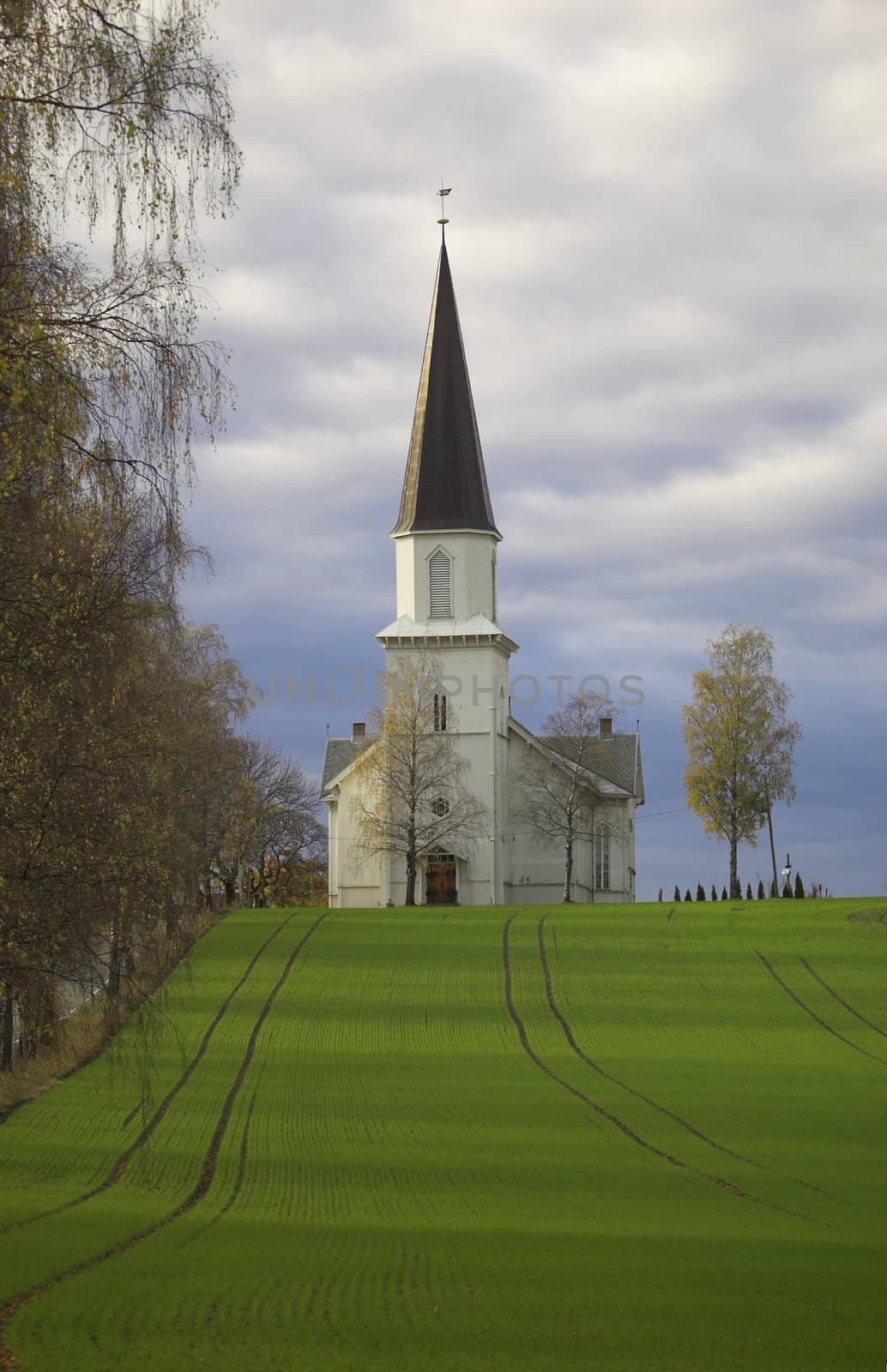 A wooden church staying on a hillock, overa a grass field, with an alleyway to one side. The church is situated in Fet, Norway.
