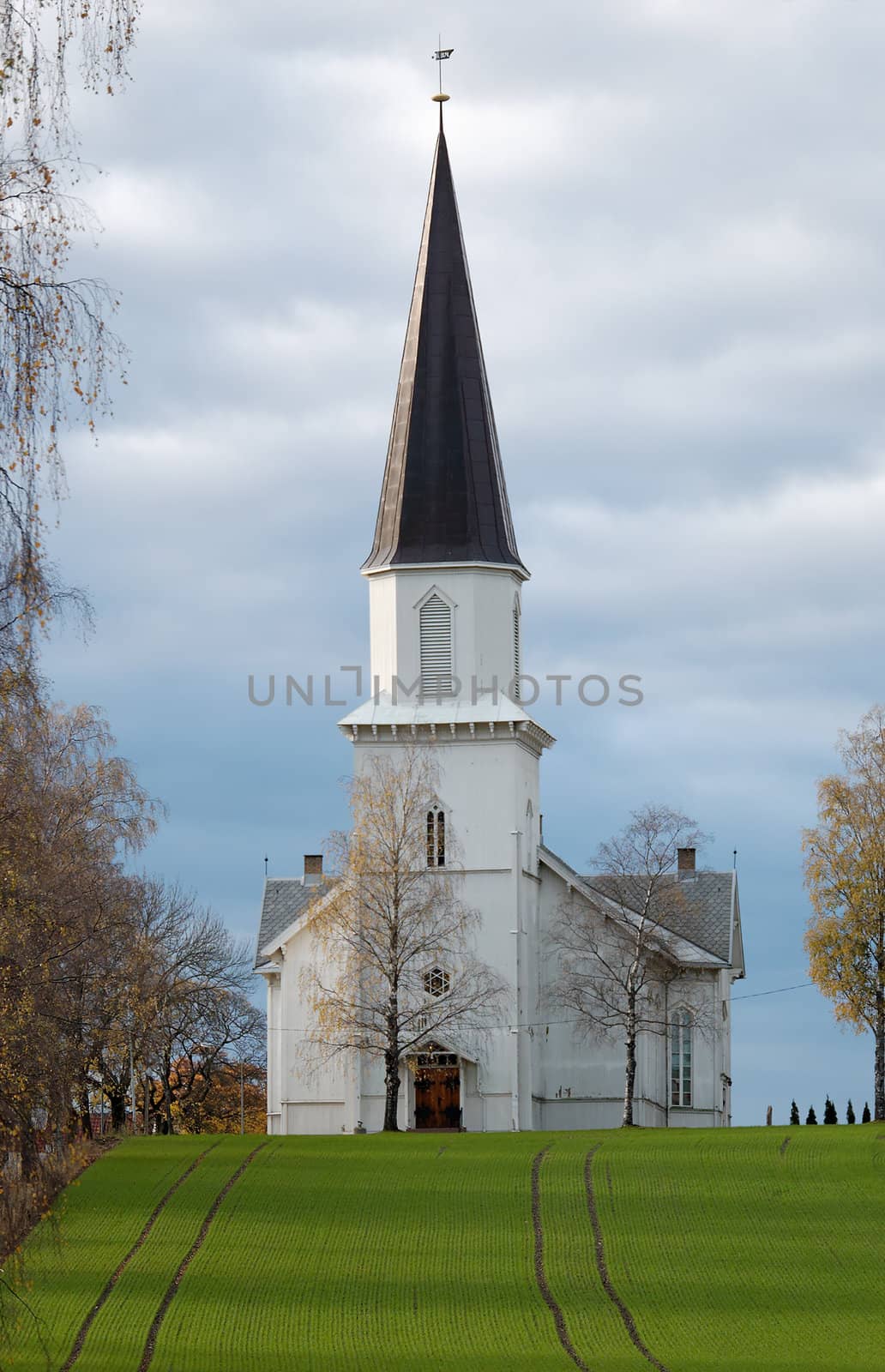 A wooden church staying on a hillock, overa a grass field, with an alleyway to one side. The church is situated in Fet, Norway.
