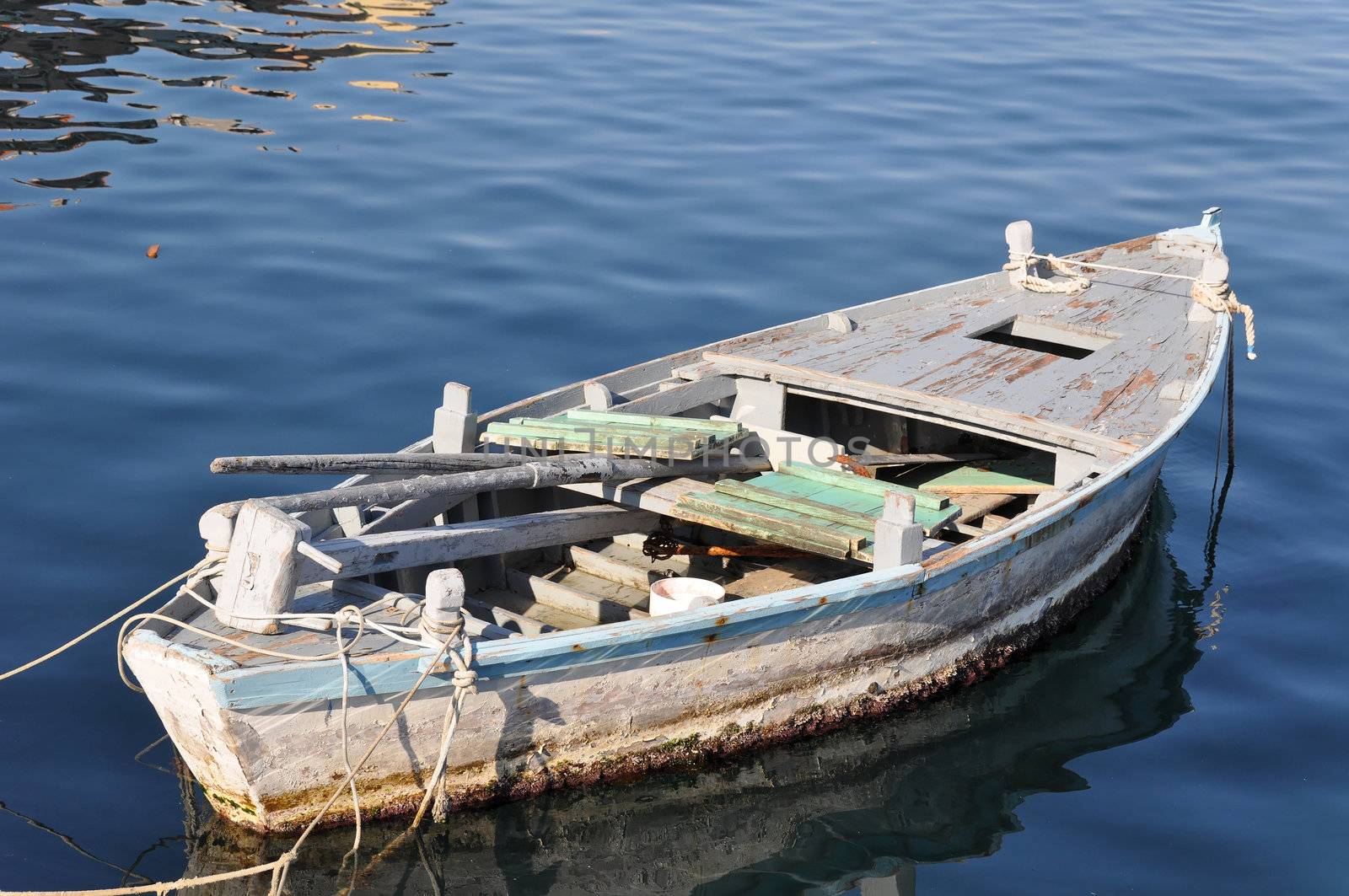 Old wooden fisher boat tied to buoys (not visible) in a Croatian harbor. Inside the boat you can see a wooden mast and paddles plus fishing equipment. The water has a great blue tone and some reflection on tiny waves.