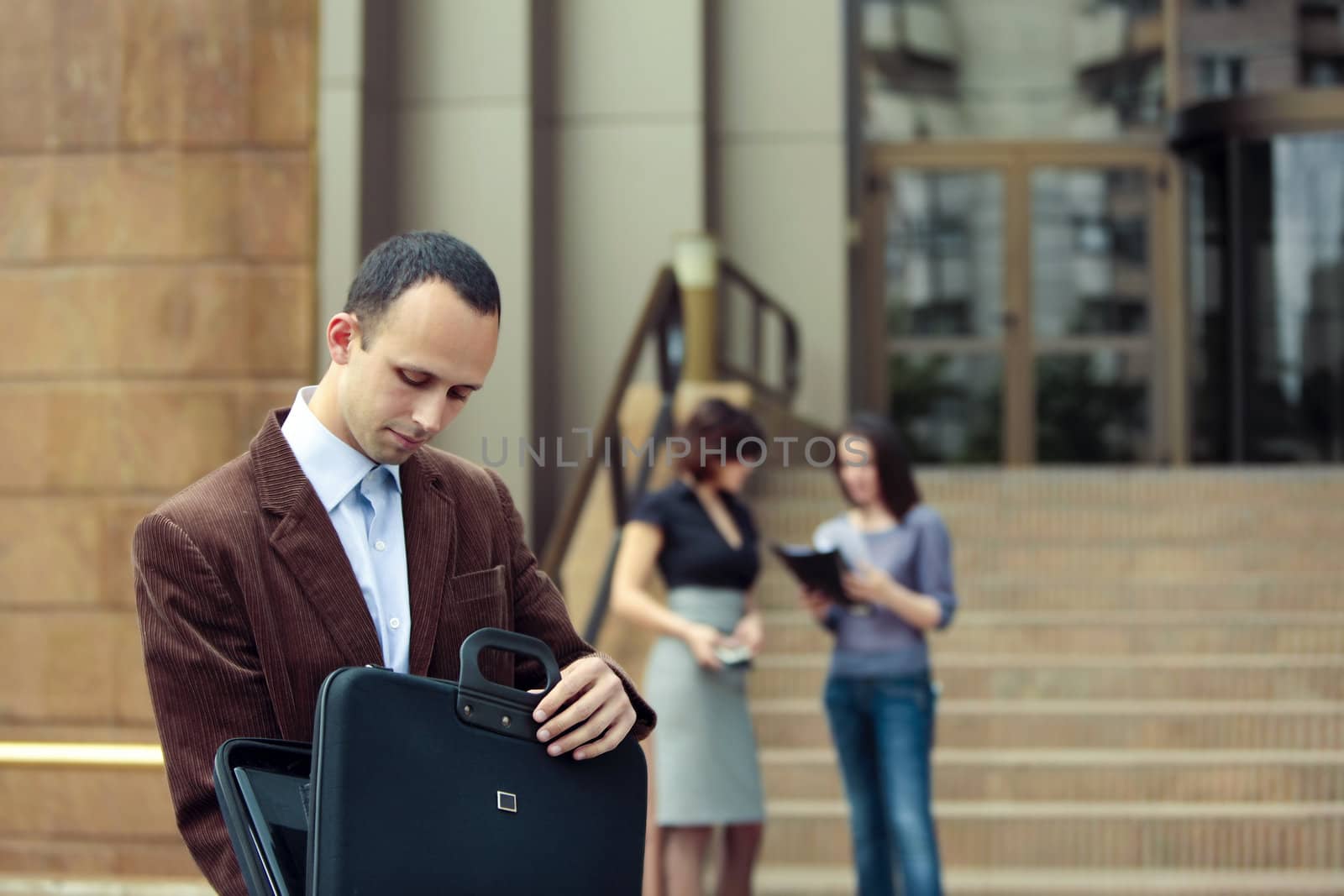 business man looking in his briefcase and business ladies talking in the background in front of a building 