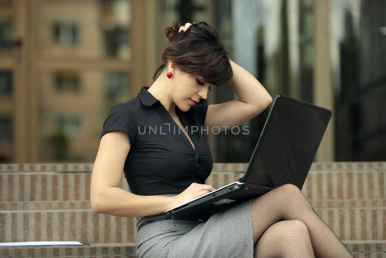 busy young attractive lady playing with her hair and looking at a laptop on the stairs in front of an office building