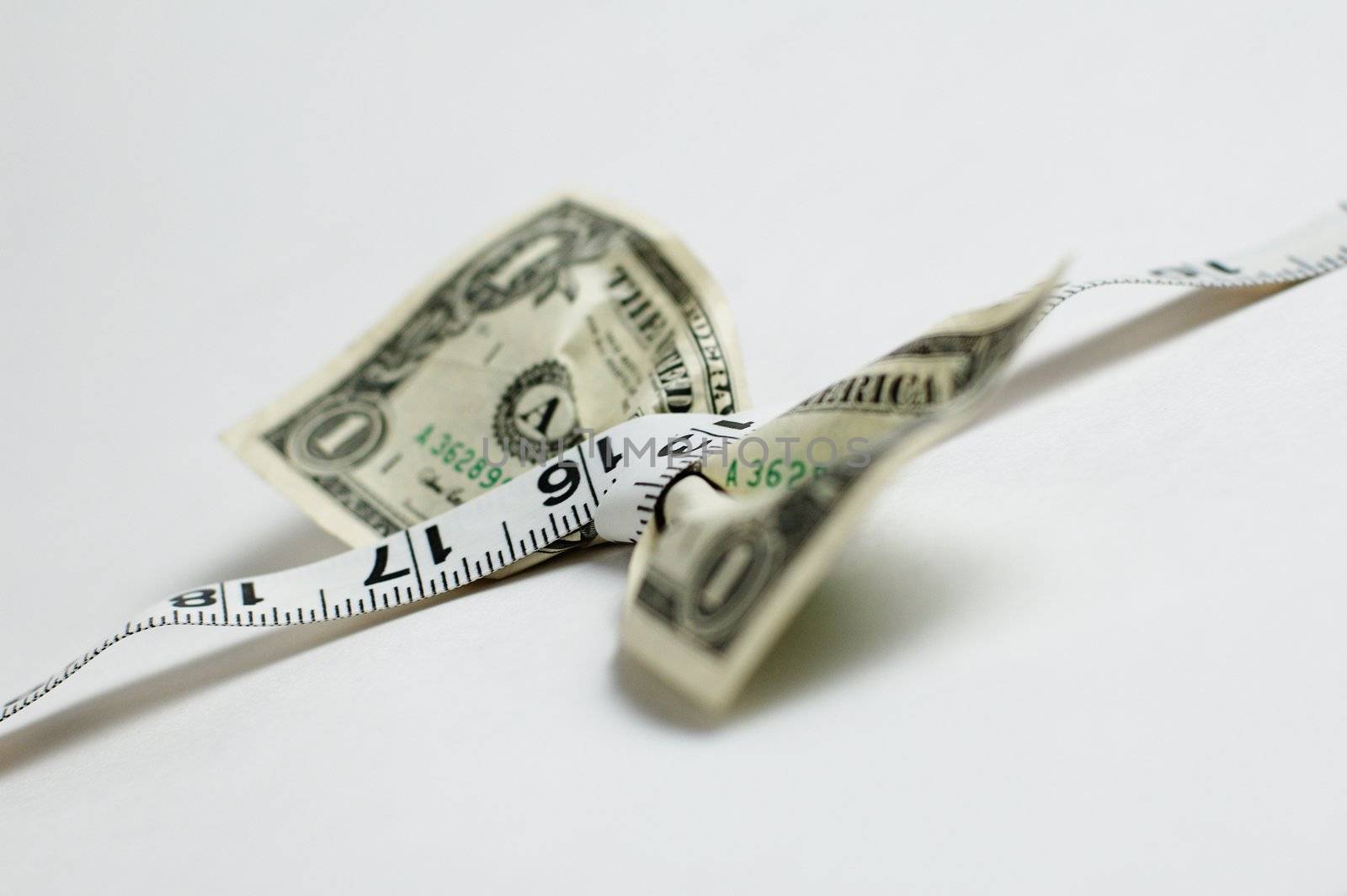 Dollar bill being squeezed by tape measure on white background.