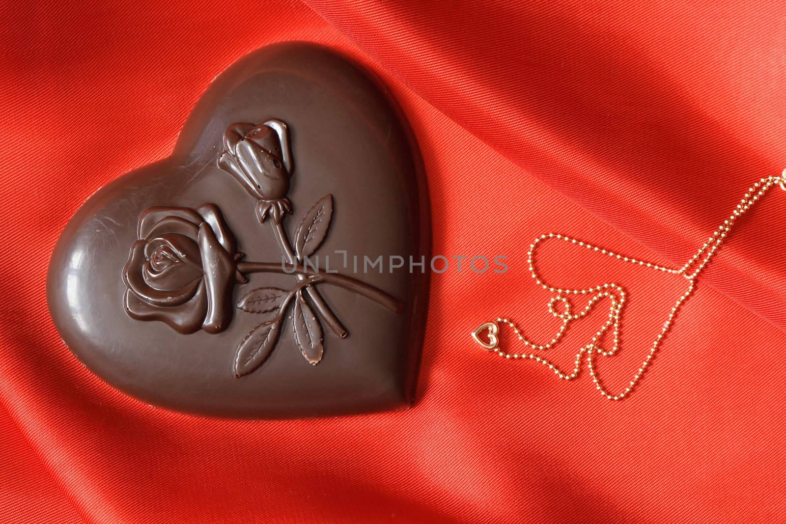 Closeup of chocolate heart and gold necklace lying on red silk textile with copy space