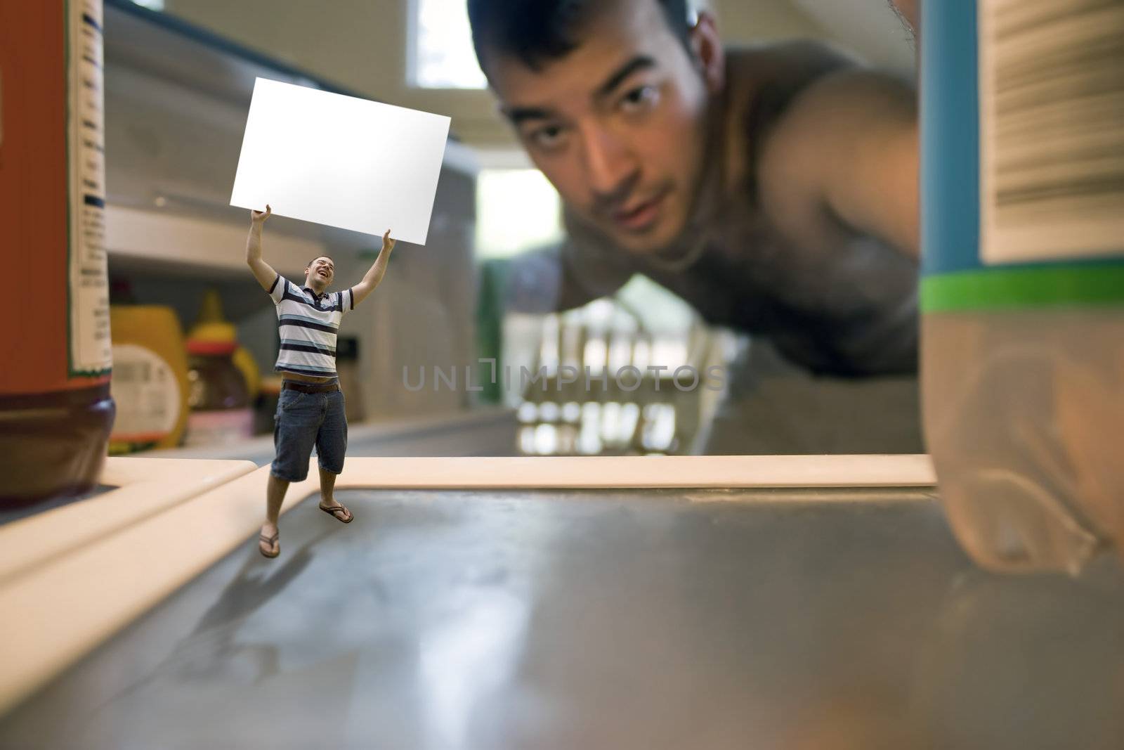 A miniature man holding up a blank sign inside the refrigerator.  Possible text on the sign could be WHATS FOR DINNER or WE NEED FOOD.  Use your imagination!  Shallow depth of field.
