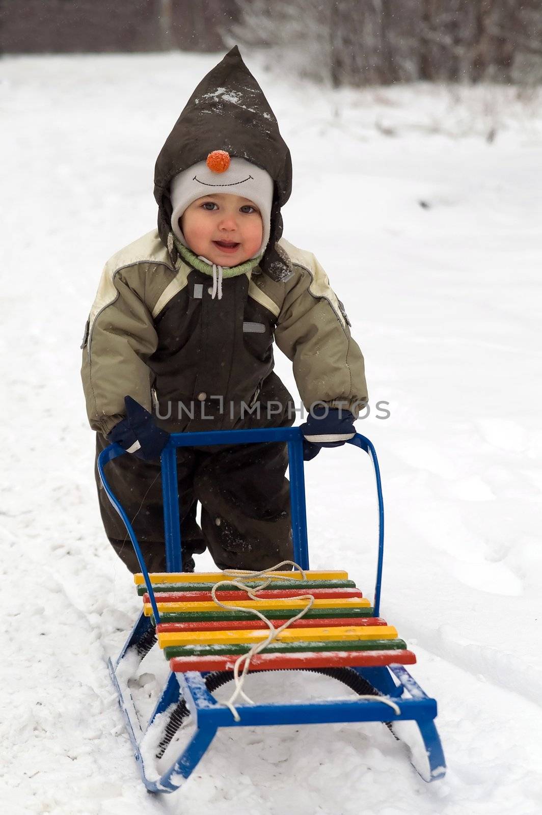 Baby in green coat pushing sled in snow-covered park