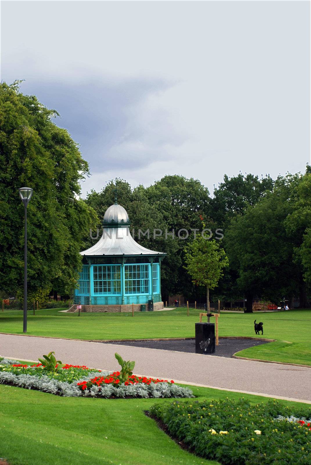 A park's green space with trees and grass, with a bandstand and a dog running with a ball.