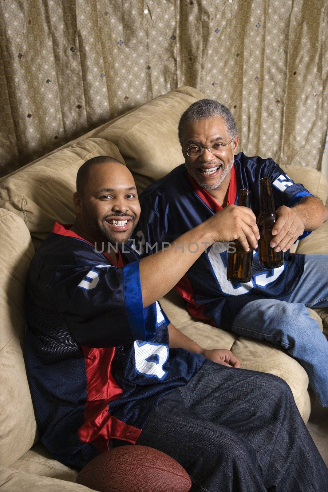 Portrait of an African-American father and son toasting with beer on couch.