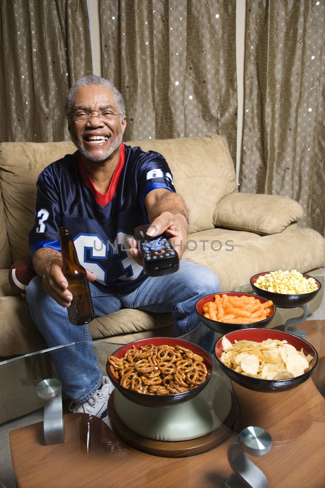 Portrait of a Middle-aged African-American man wearing a football jersey surrounded by snacks pointing a remote at viewer.