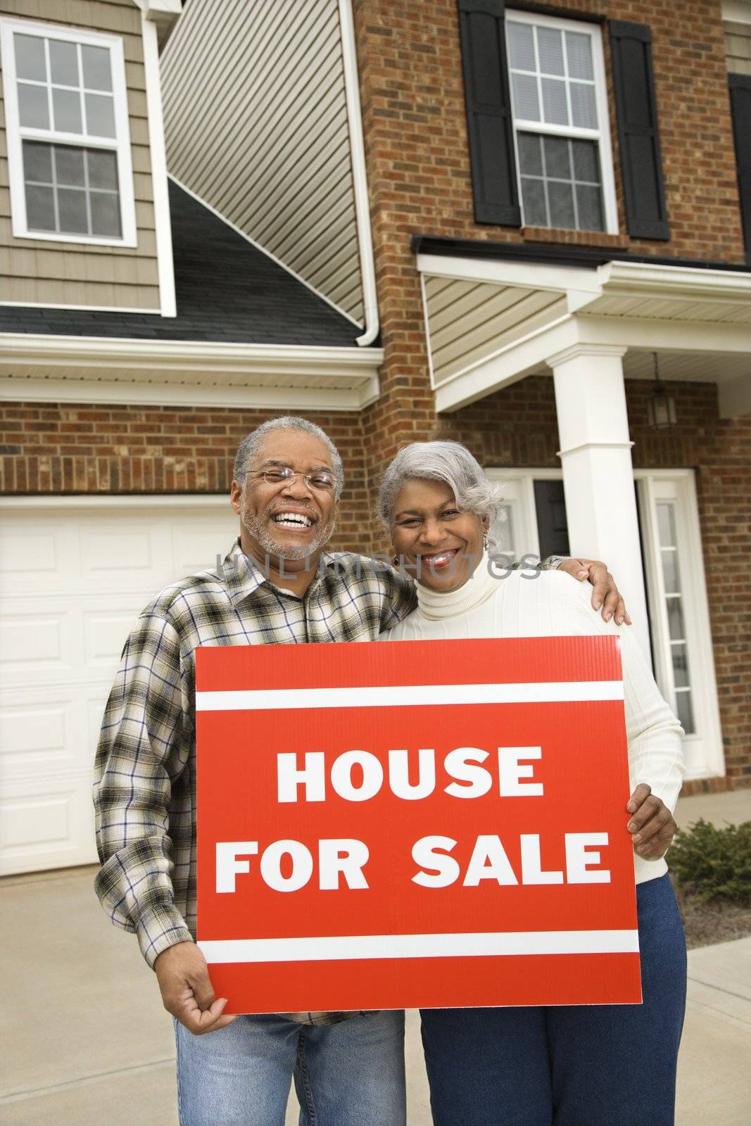 Portrait of middle-aged African-American couple outside house with for sale sign.