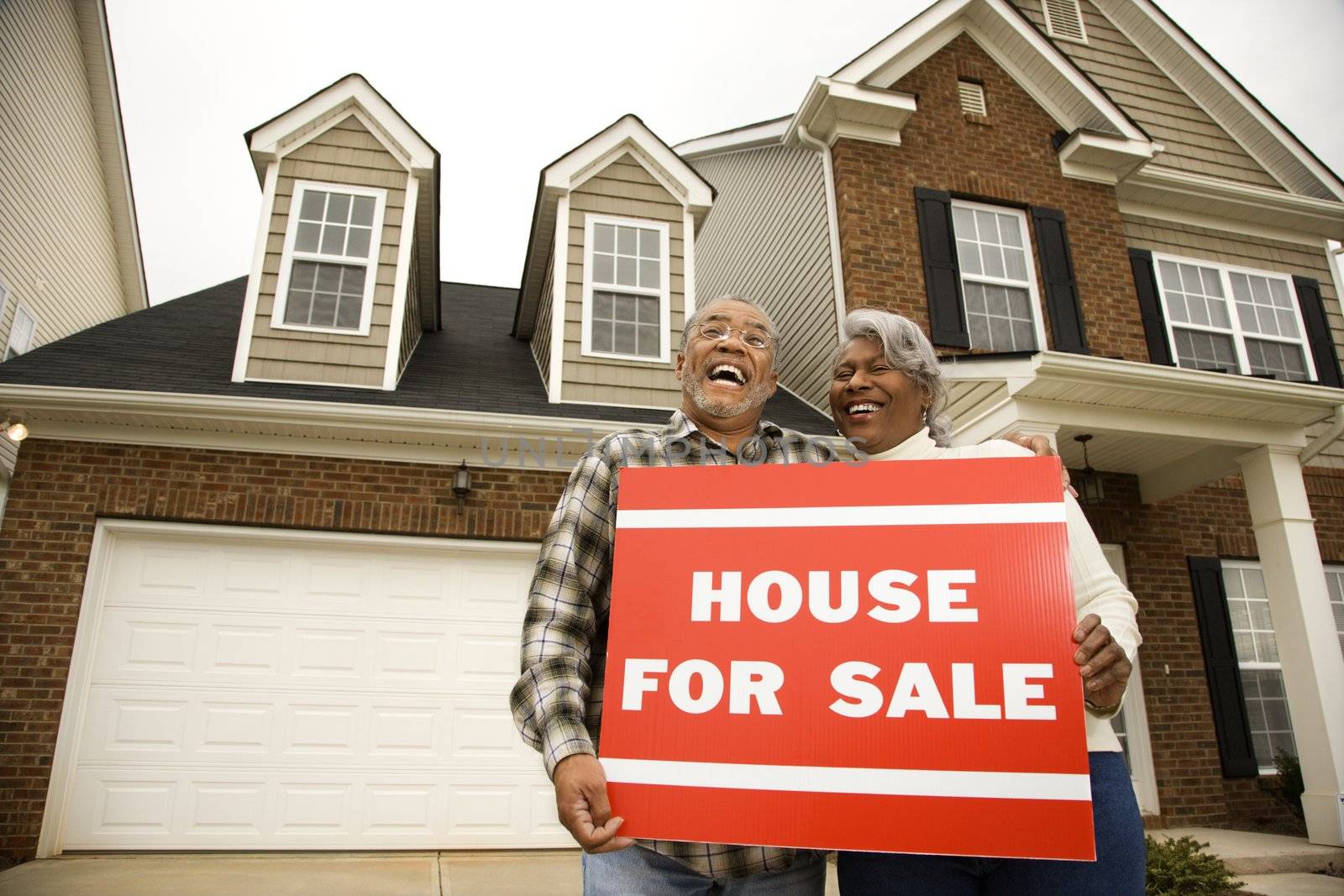 Portrait of middle-aged African-American couple outside house with for sale sign.