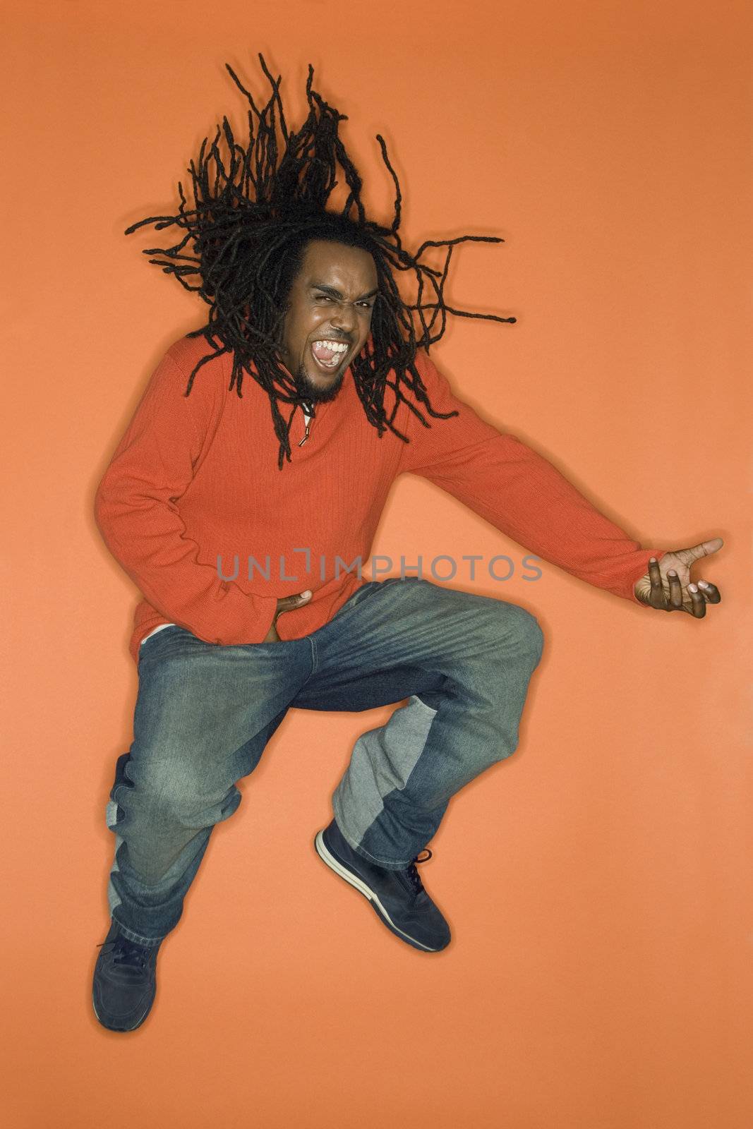 African-American mid-adult man on orange background jumping while playing air guitar.