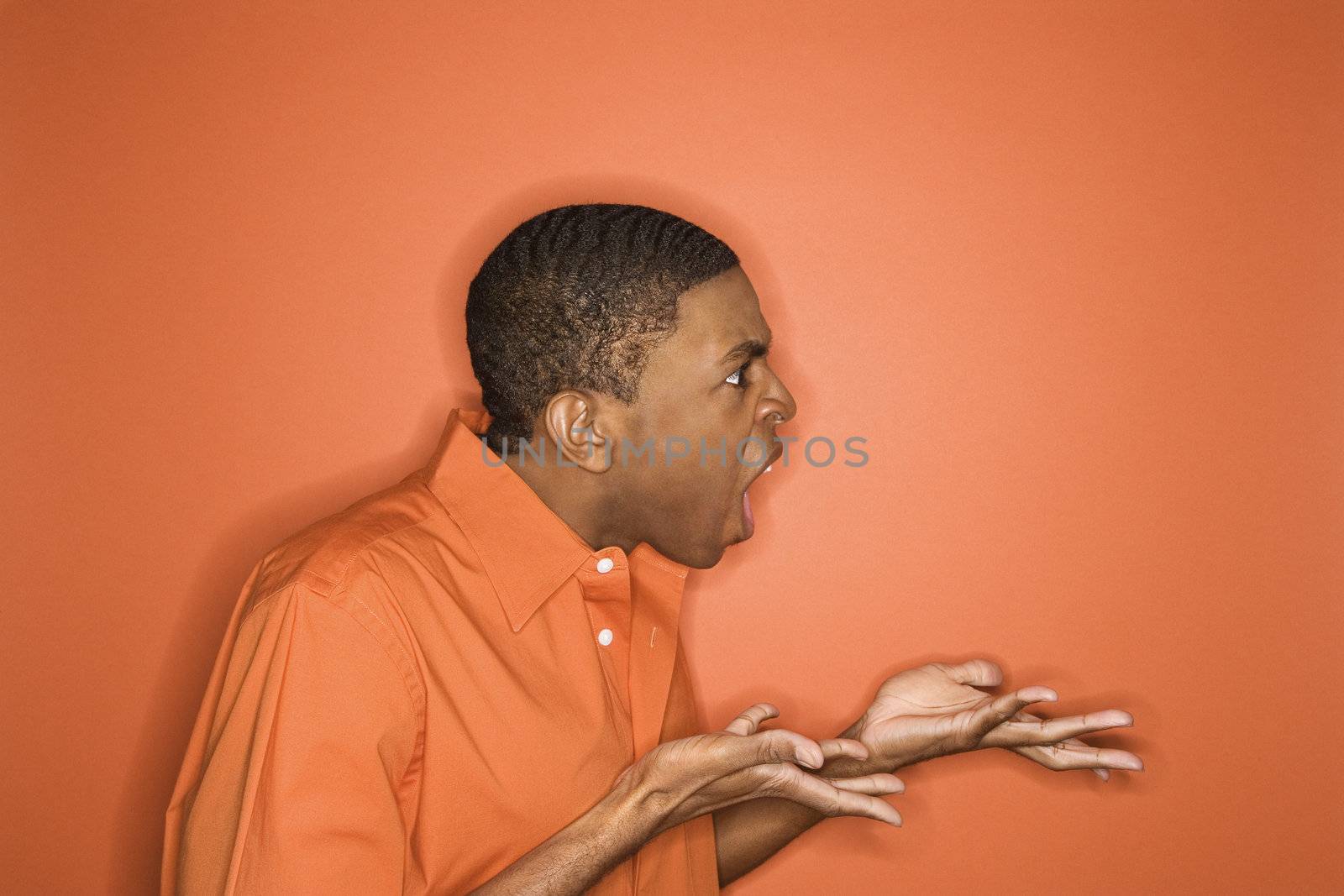 Side view of young African-American man on orange background expressing anger towards unseen person.