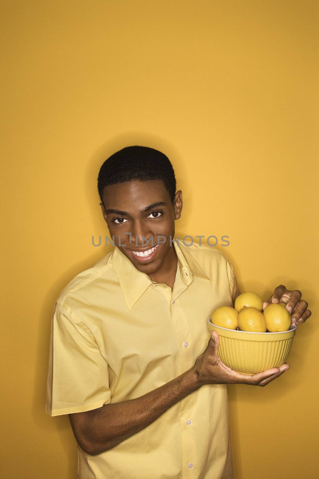 Smiling young African-American man holding bowl of lemons on yellow background.