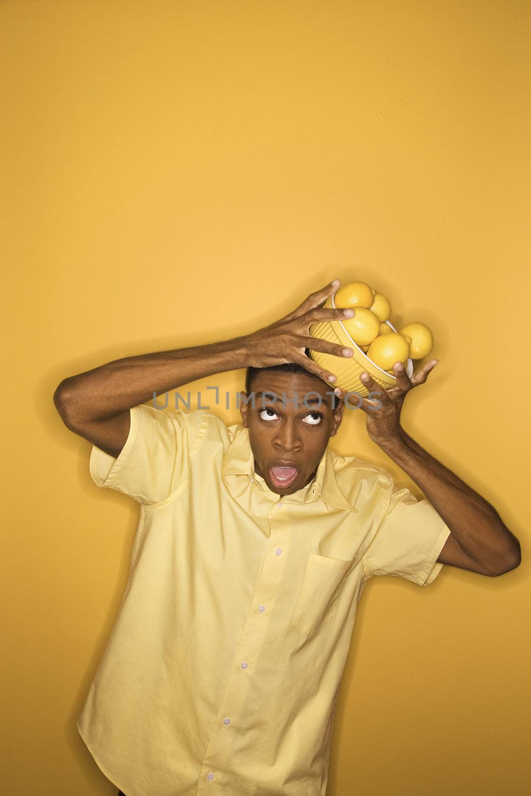 Young African-American man dropping a bowl of lemons balancing on his head, on yellow background.