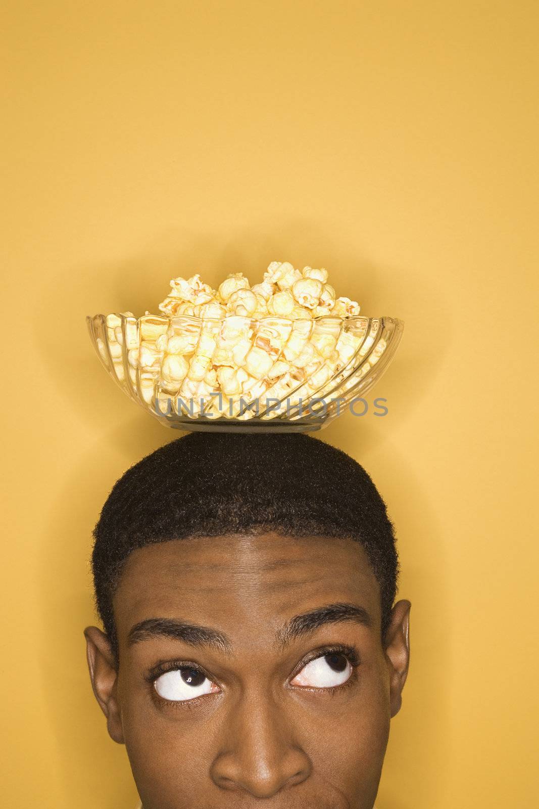 Young African-American man balancing bowl of popcorn on his head on yellow background.