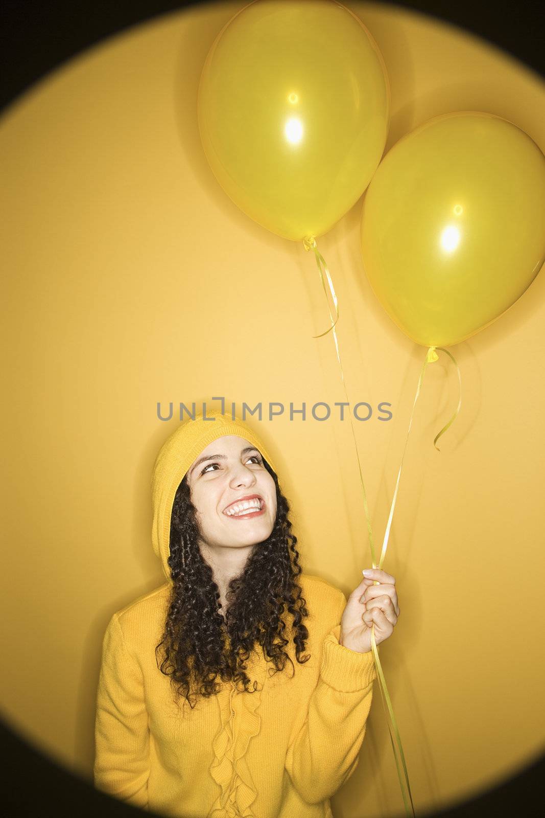 Smiling young Caucasian woman with balloons wearing yellow raincoat on yellow background.