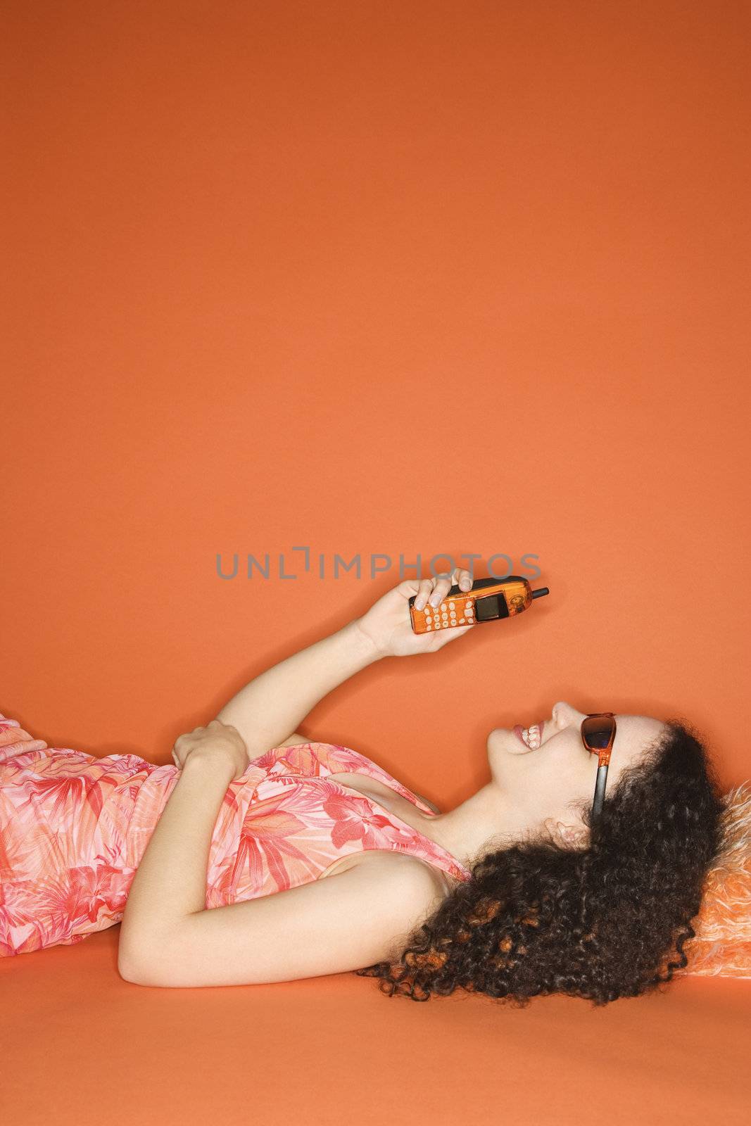 Young Caucasian woman lying on floor looking at cell phone on orange background.