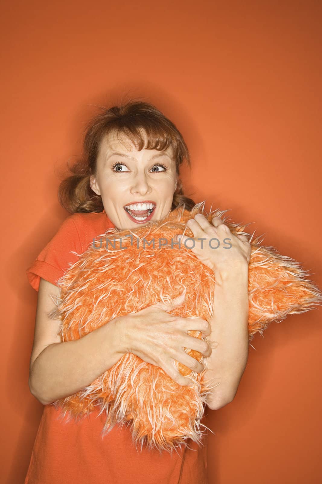 Caucasian mid-adult woman on orange background hugging fuzzy orange pillow.