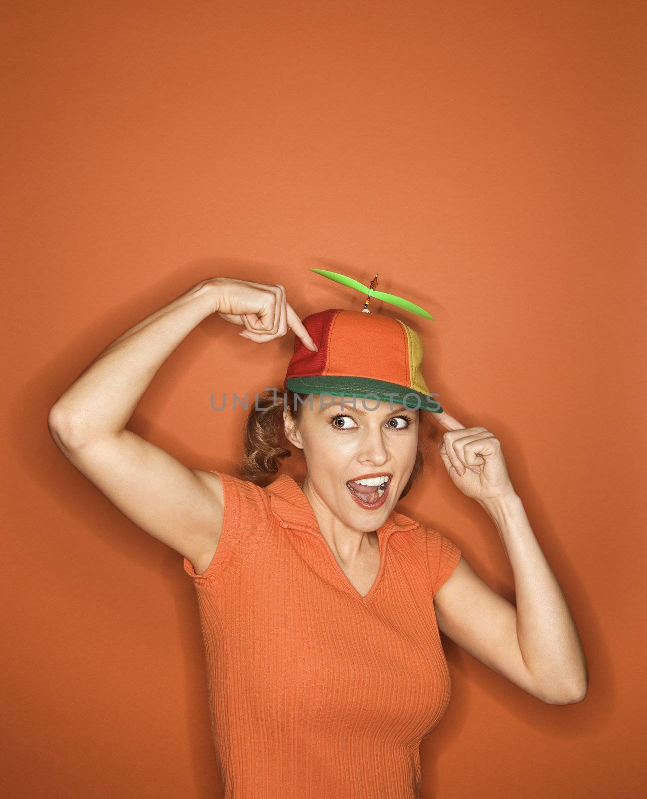 Smiling Caucasian mid-adult woman pointing and wearing propeller cap on orange background.