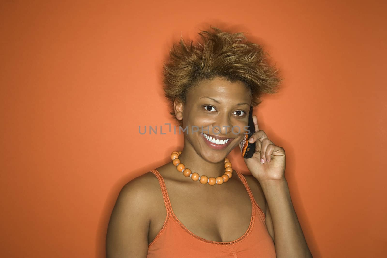Portrait of smiling young African American adult woman on orange background talking on cellphone.