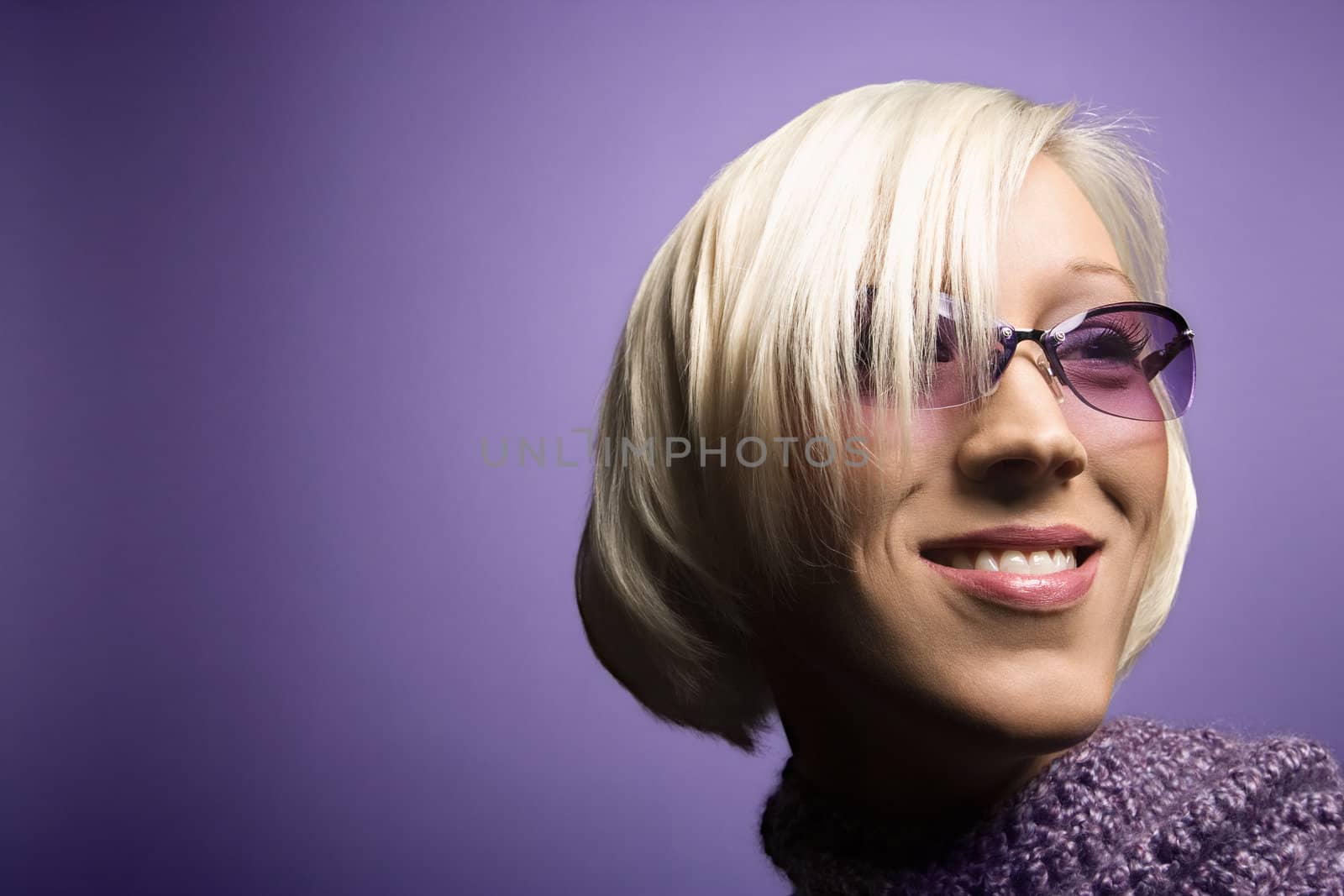 Portrait of smiling young adult Caucasian blond woman on purple background wearing sunglasses and scarf.