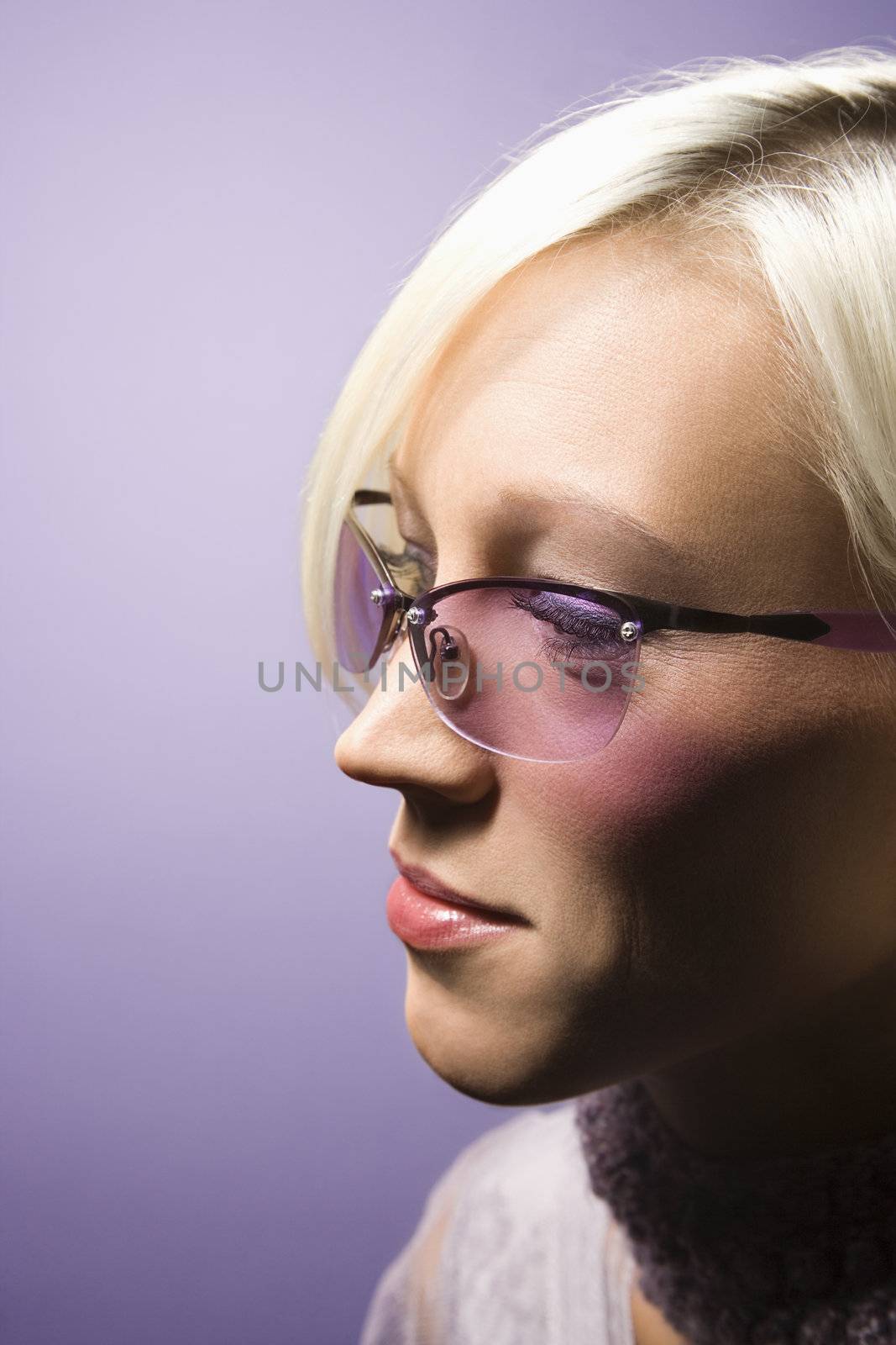 Close-up side view portrait of young adult Caucasian woman on purple background wearing sunglasses.
