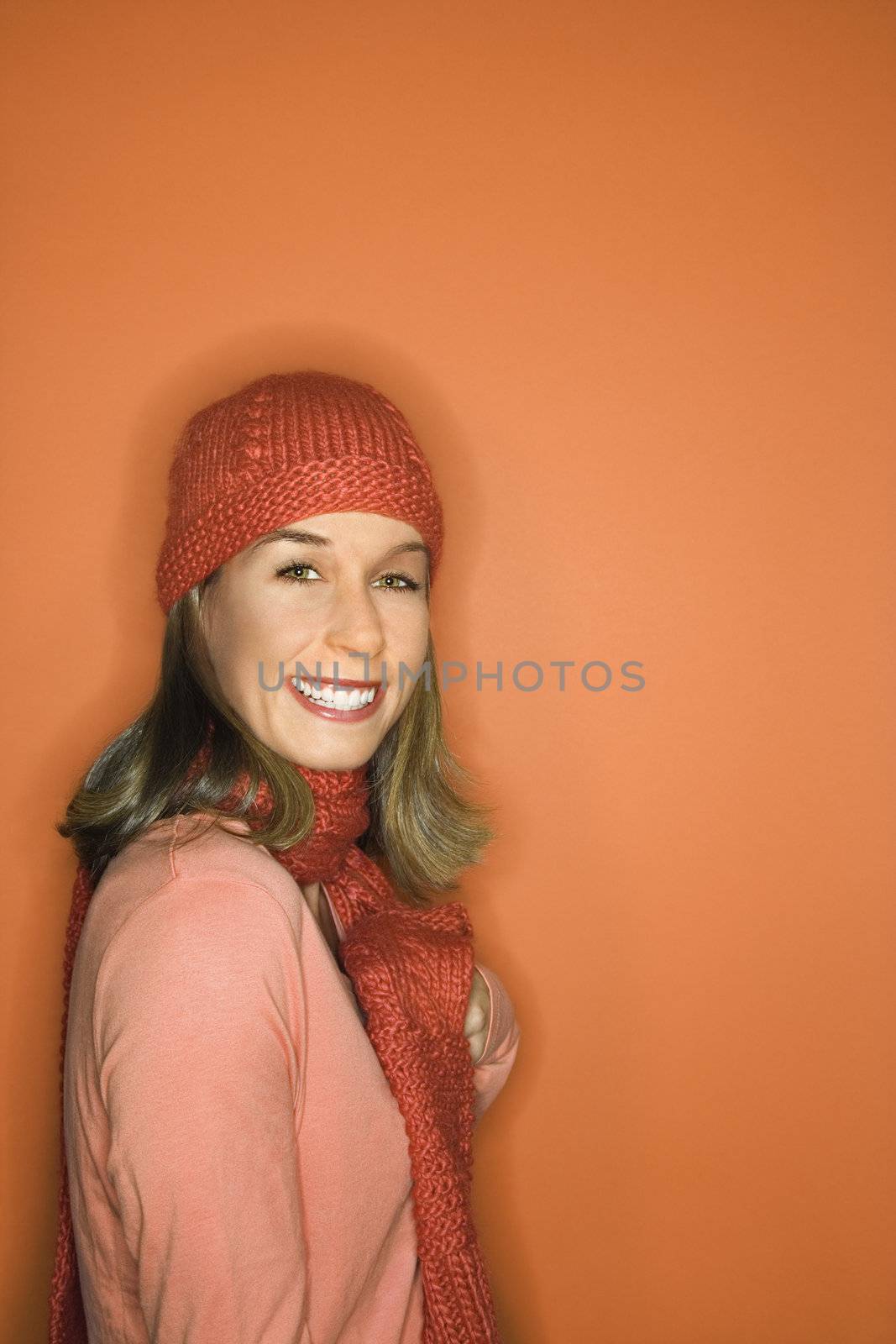 Portrait of smiling young adult Caucasian woman on orange background wearing winter hat and scarf.
