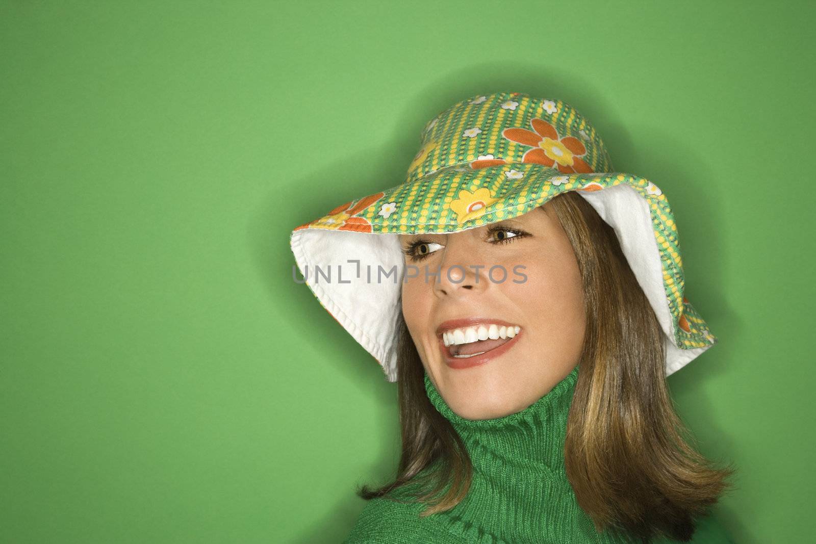 Portrait of smiling young adult Caucasian woman on green background wearing floppy hat.
