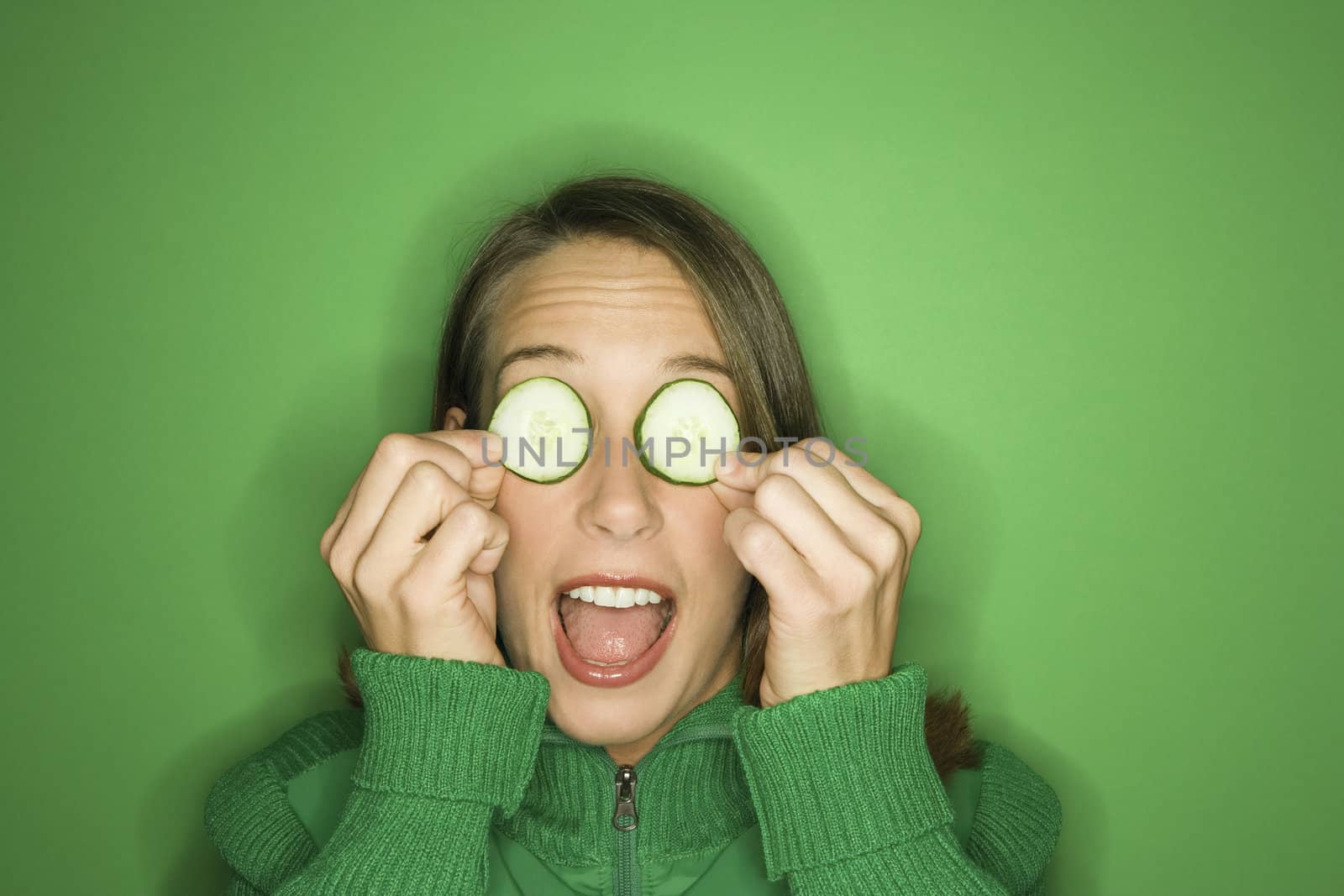 Portrait of young adult Caucasian woman on green background holding cucumber slices over her eyes.