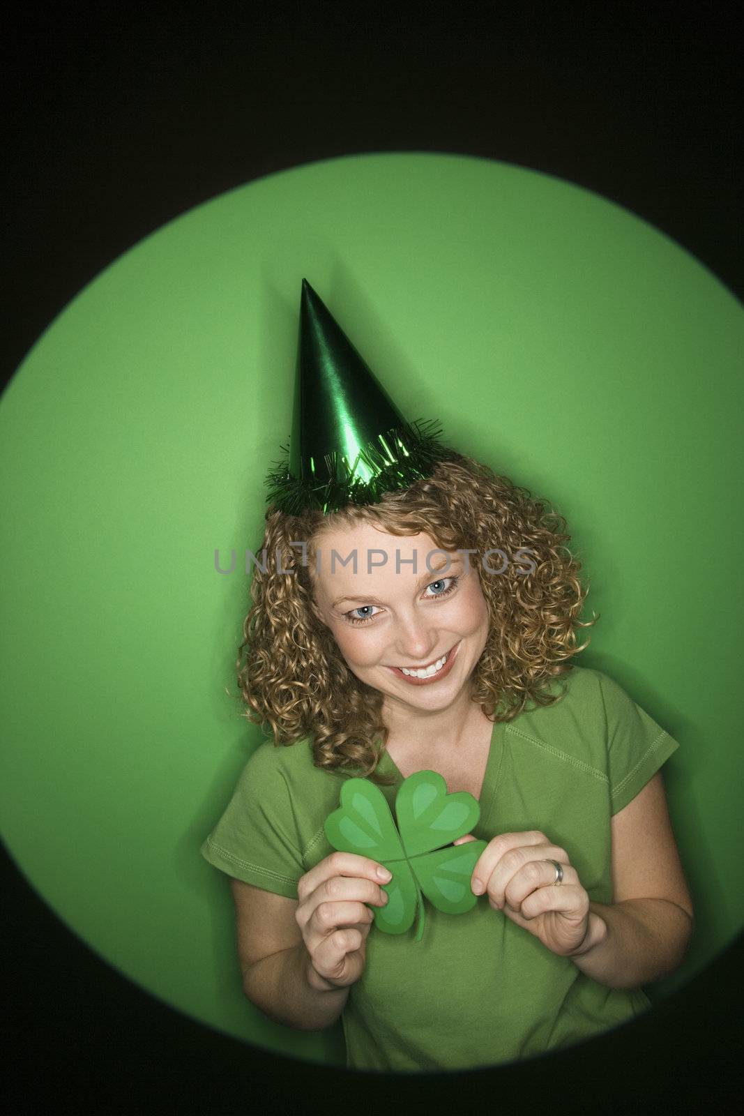 Vignette of smiling young adult Caucasian woman on green background wearing Saint Patricks Day hat and holding shamrock.
