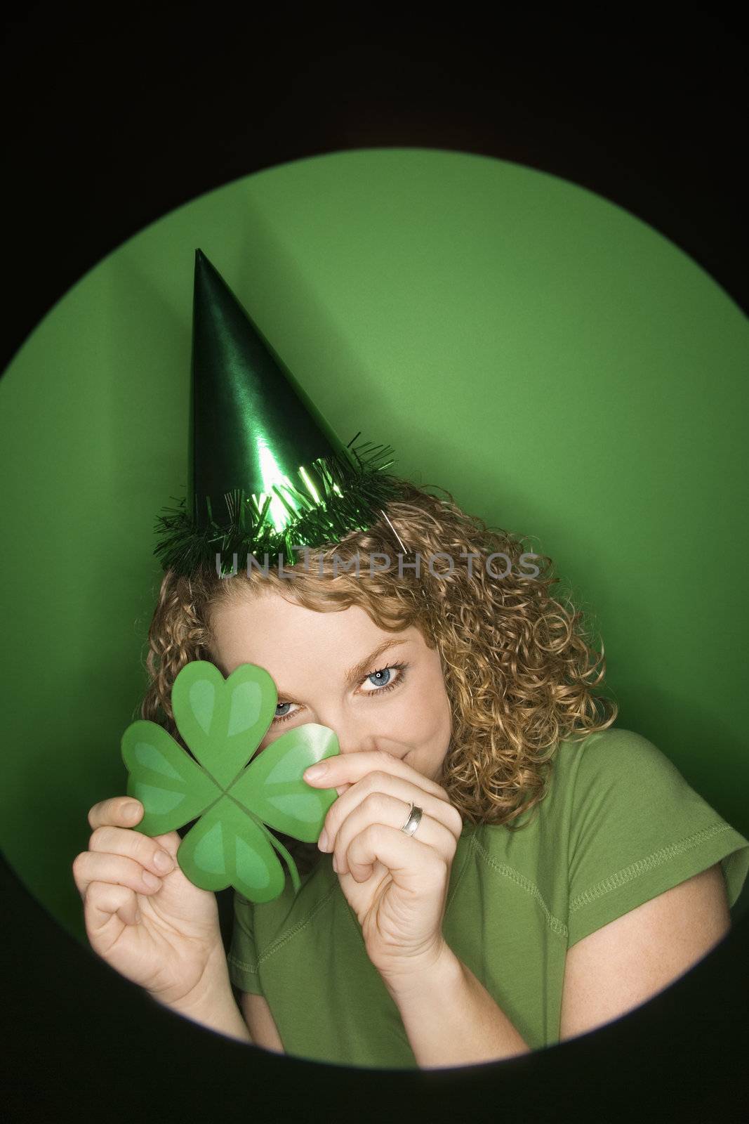 Vignette of young adult Caucasian woman on green background wearing Saint Patricks Day hat and holding shamrock.