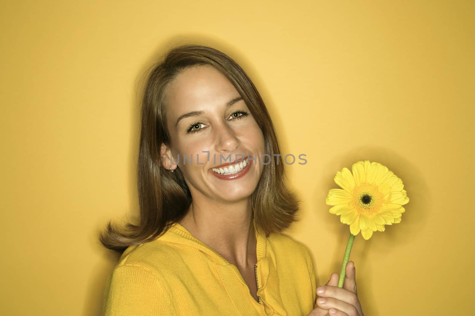 Portrait of smiling young adult Caucasian woman on yellow background holding flower.