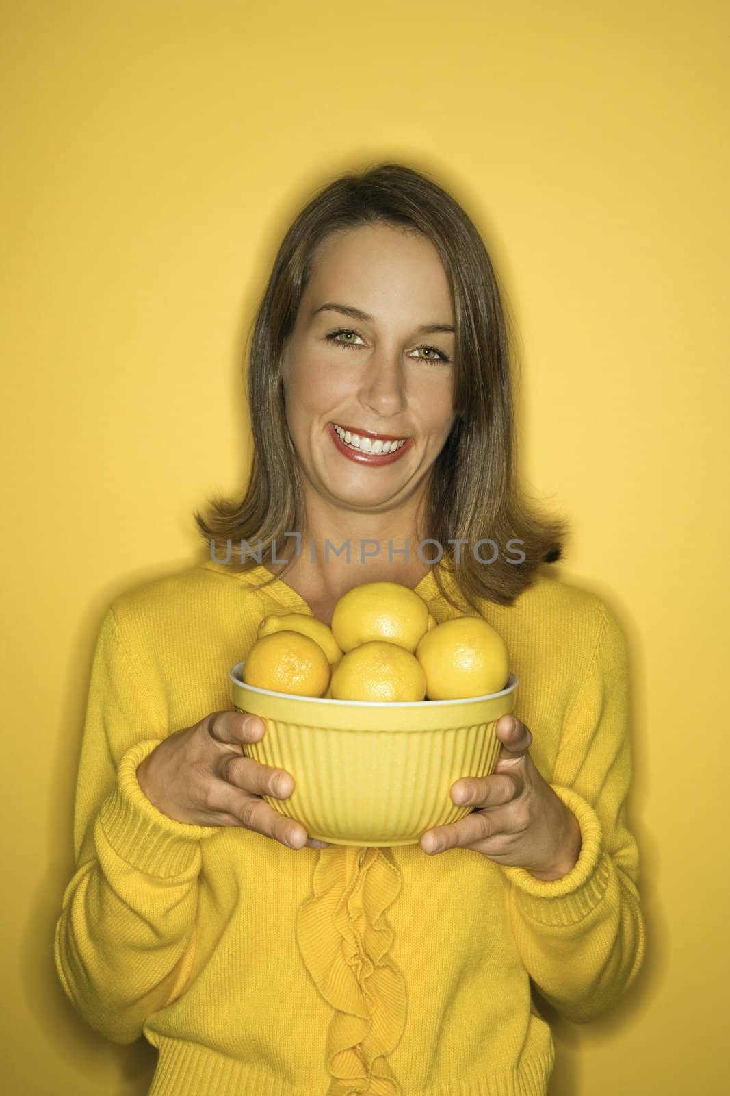 Portrait of smiling young adult Caucasian woman on yellow background holding bowl of lemons.