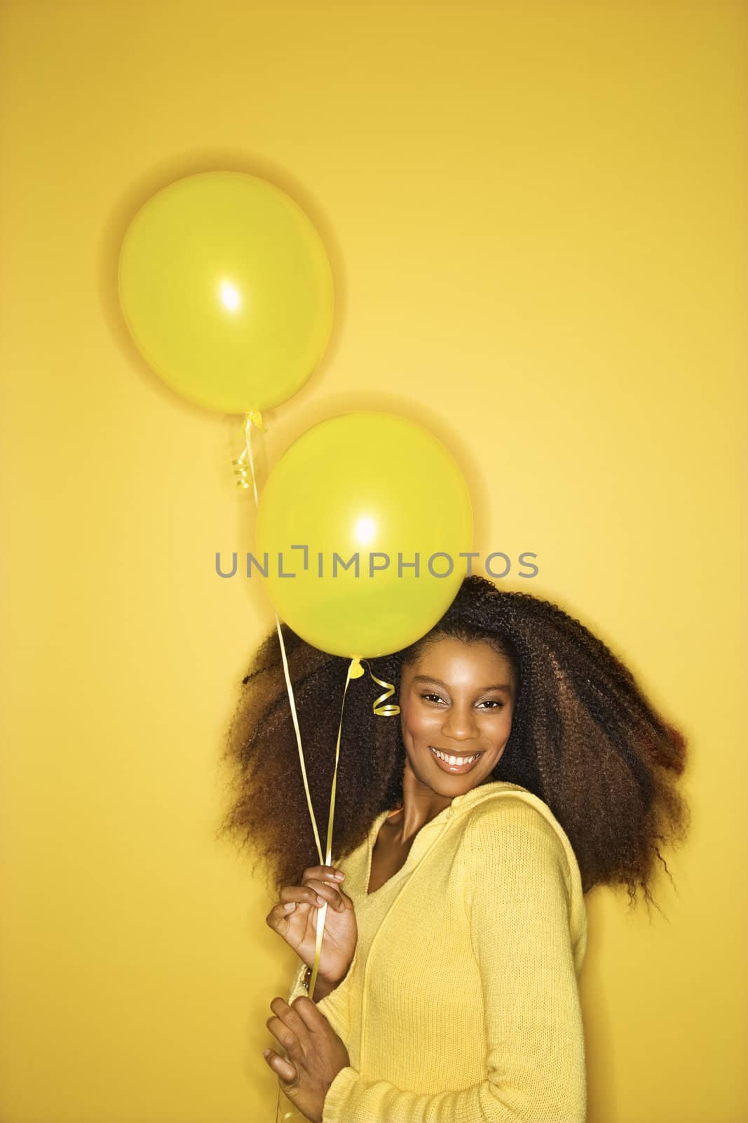 Portrait of smiling young African-American adult woman looking sweet on yellow background holding balloons.