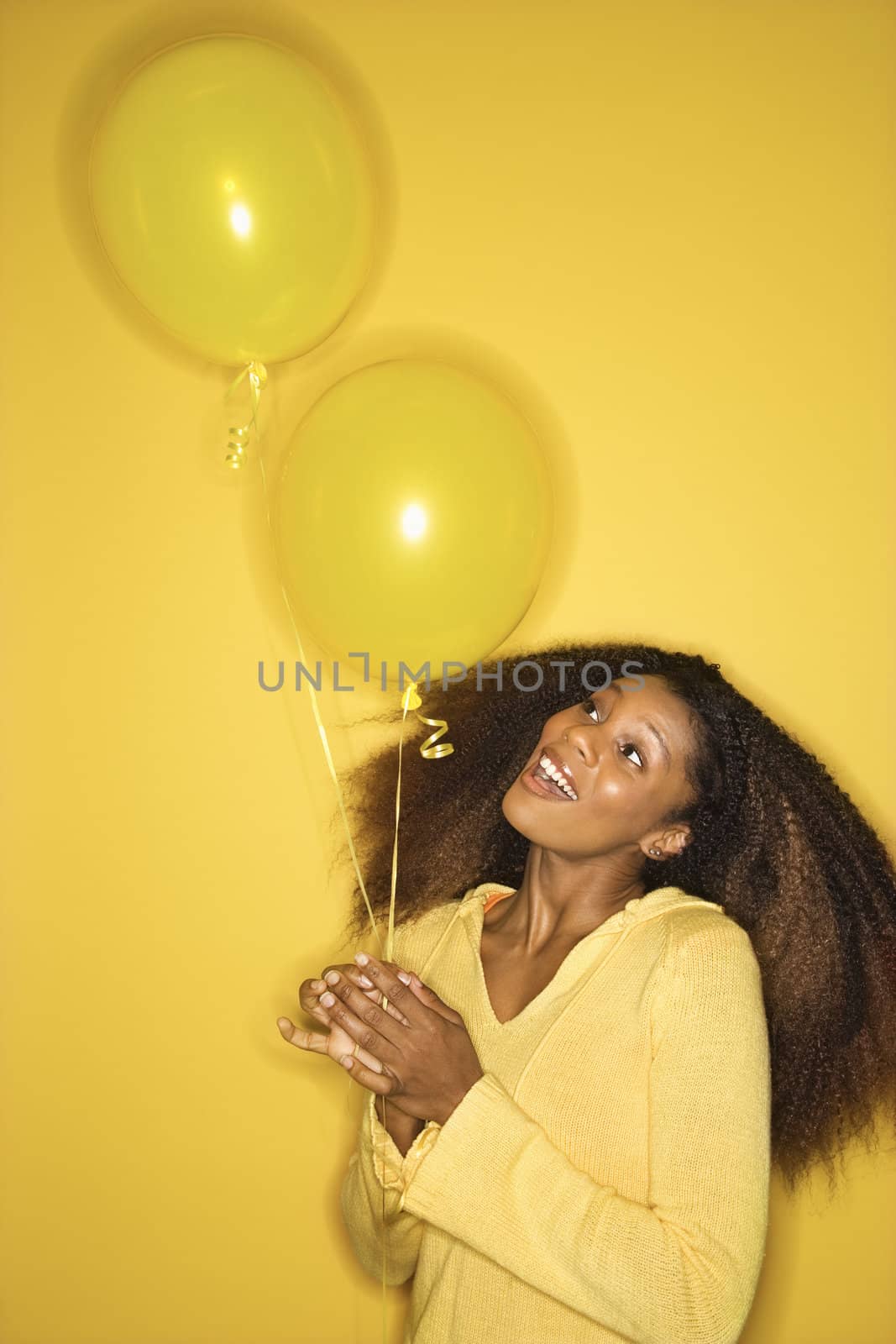 Portrait of smiling young African-American adult woman on yellow background holding balloons.